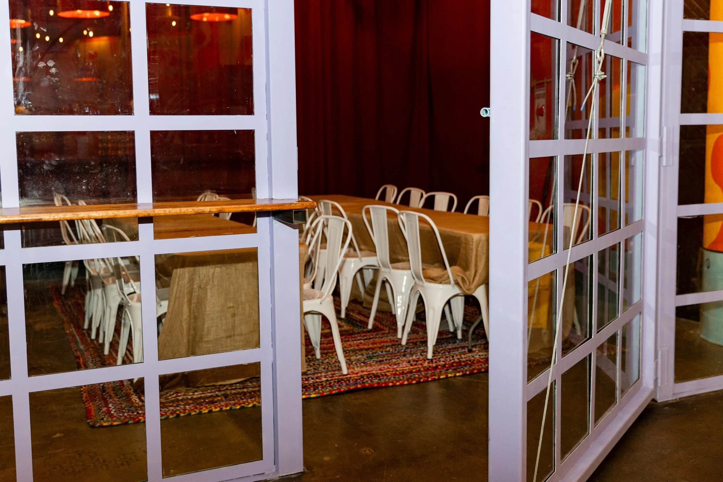 Private dining area seen through a glass partition with lavender grid frames. Inside, there are chairs around a tables able to hold large groups of up to 25 people with a cloth cover, and a colorful rug on a floor.