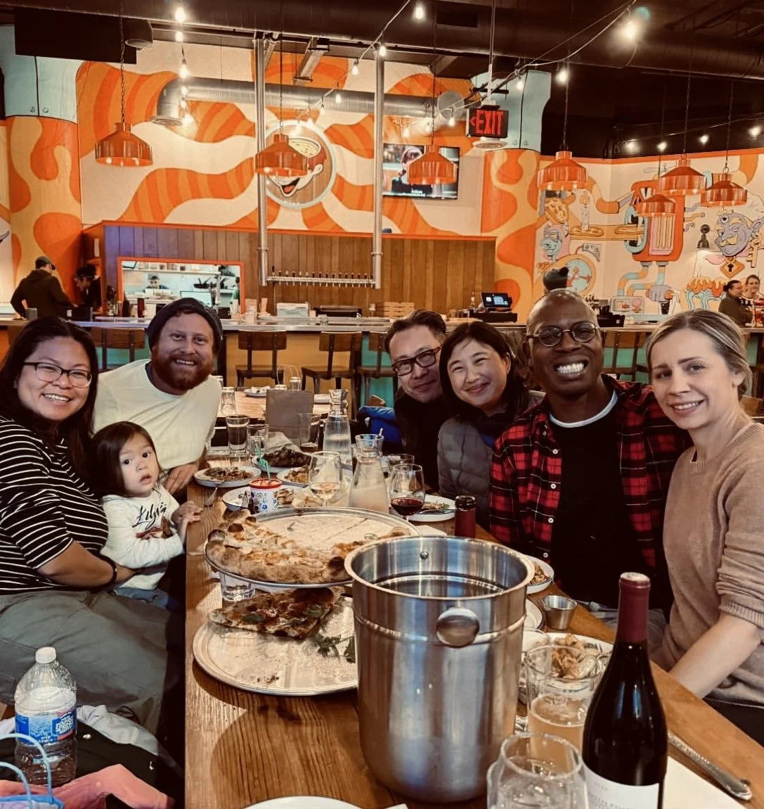 A group of seven diverse people, including a child, sitting at a long table in a colorful restaurant with orange and white walls and cartoon murals, enjoying pizza and drinks.