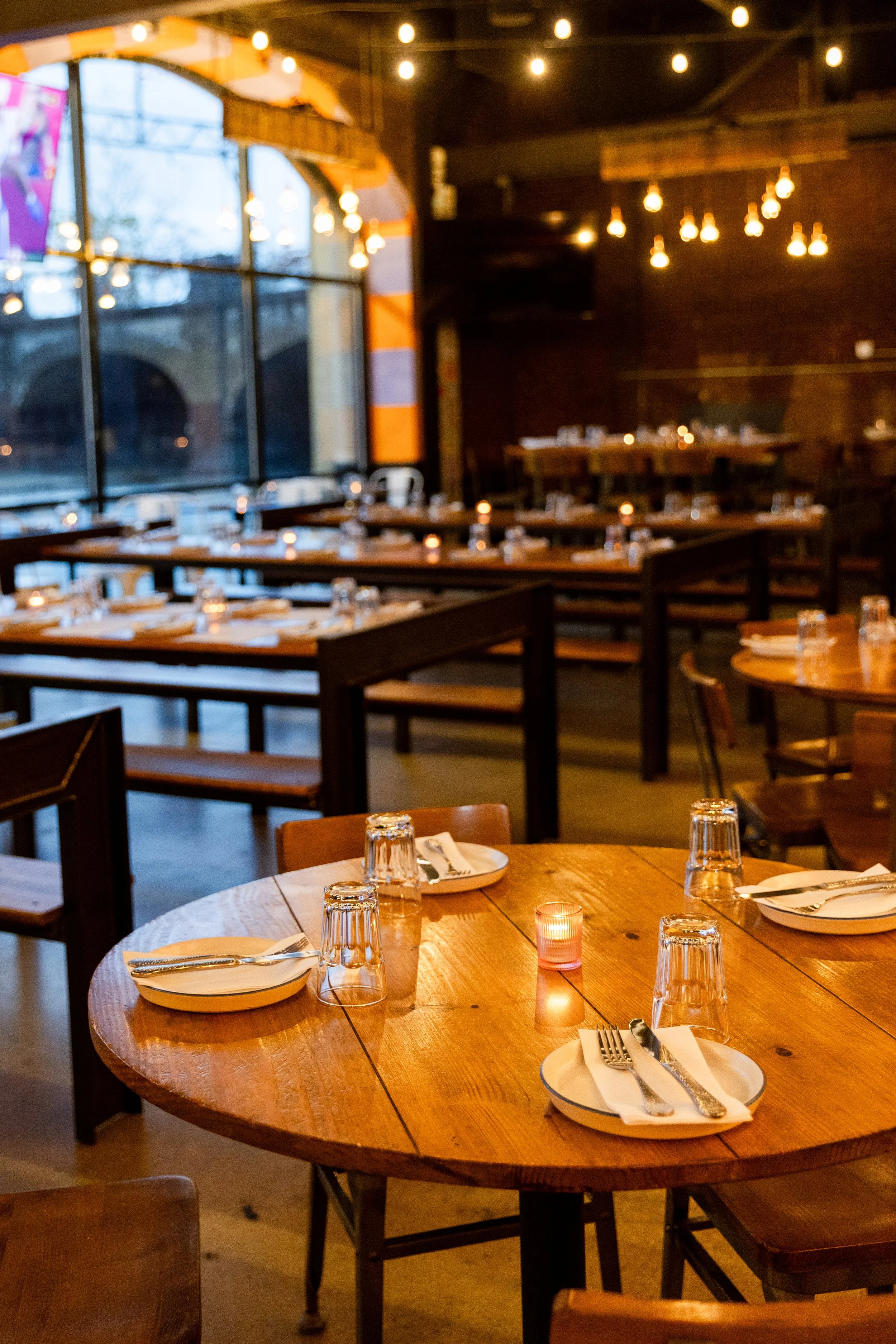 Empty restaurant with wooden tables set with plates, forks, knives, water glasses, and small candles, illuminated by warm string lights indoors.