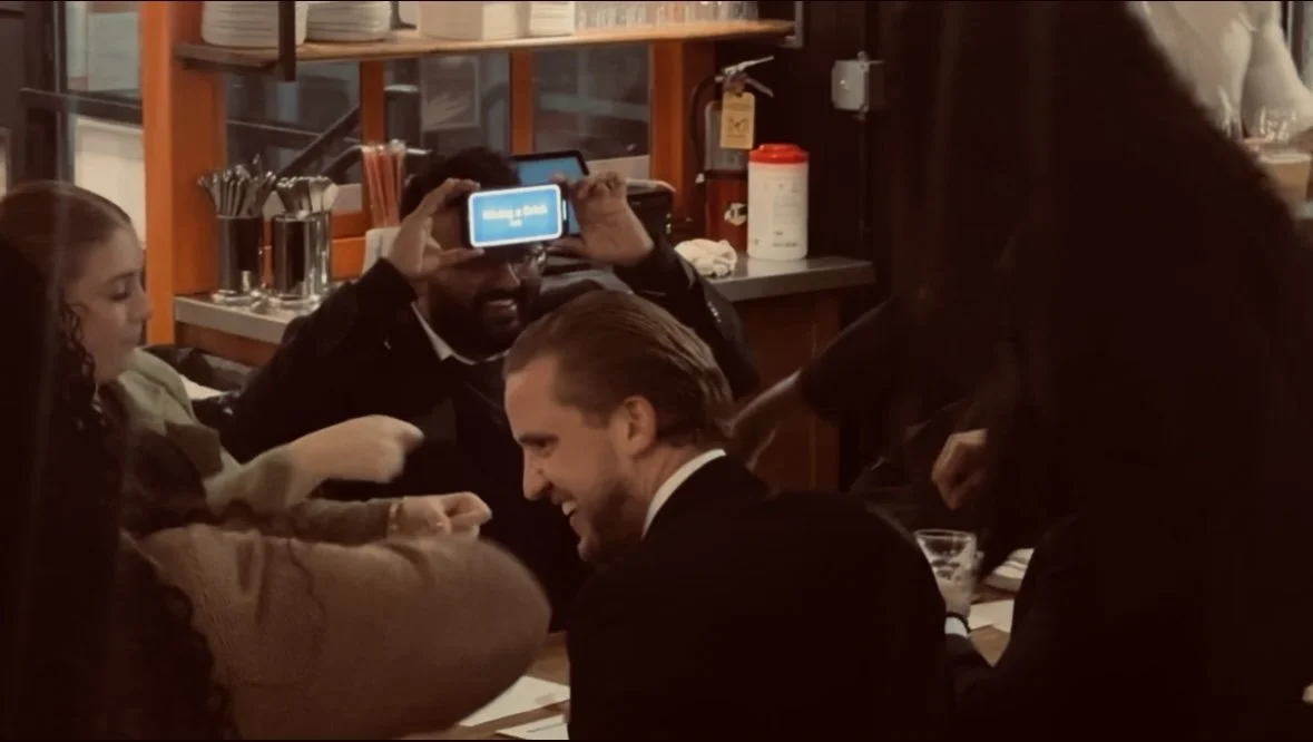 People sitting at a table in a restaurant, talking, and smiling. One person is holding a phone, capturing a photo or video.