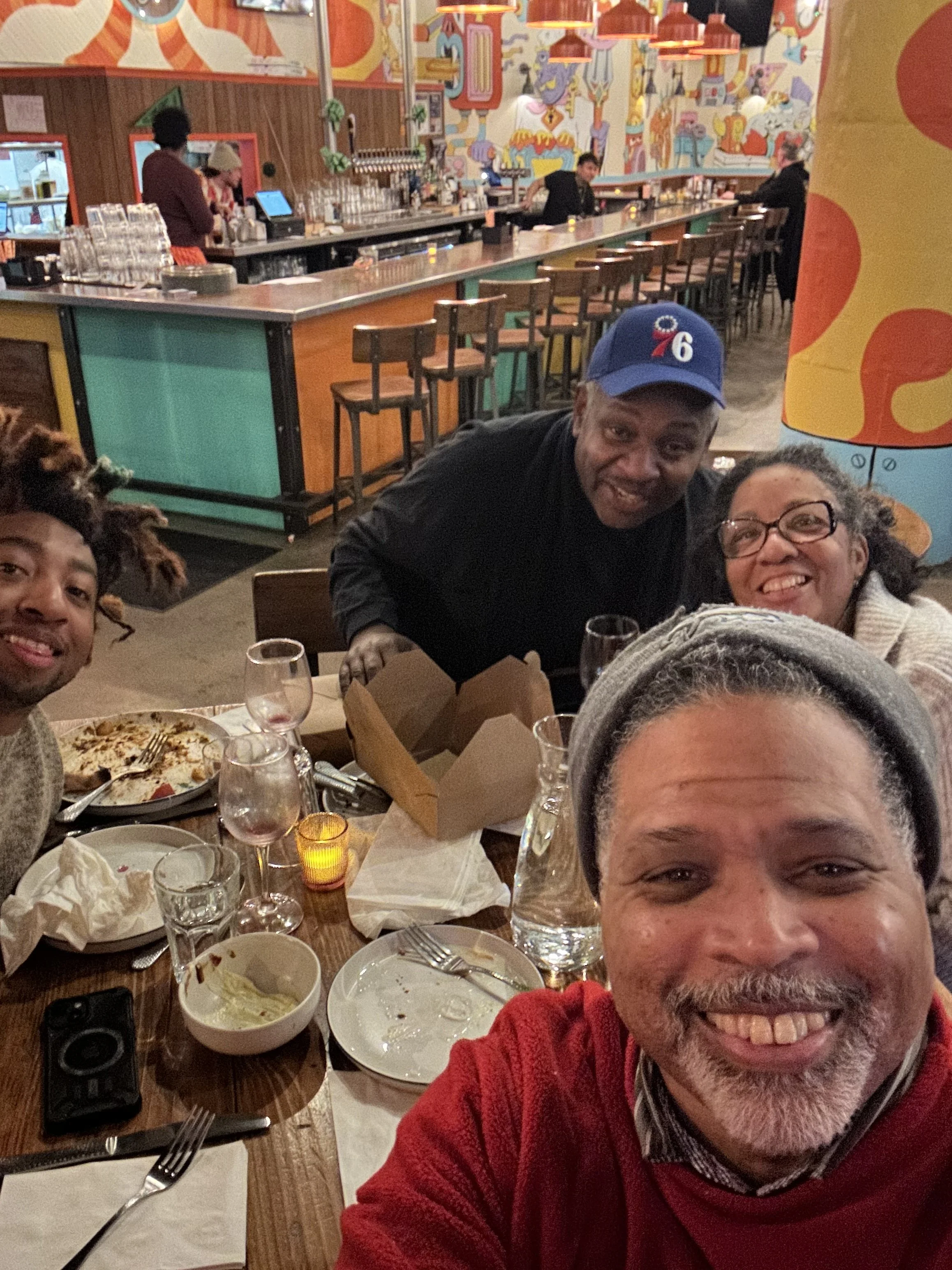 A group of five people posing for a photo at a restaurant table with leftover food, glasses, and drinks. The background shows a colorful, playful mural and a bar area.