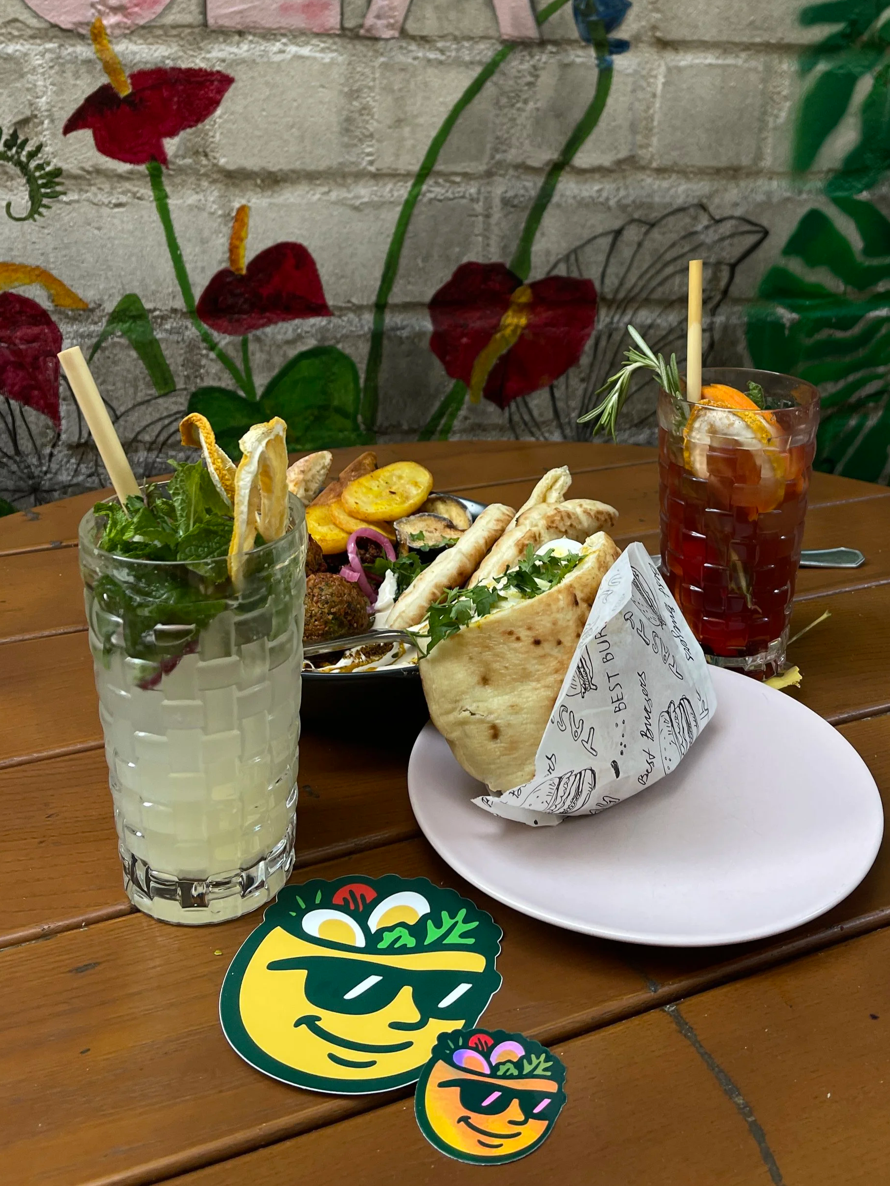 A table with two drinks, a burger wrapped in paper, and a black bowl of assorted Middle Eastern or Mediterranean food, with a painted brick wall mural in the background.