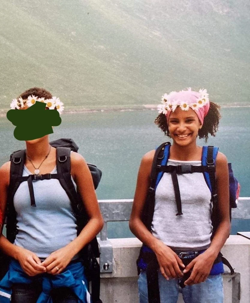Two women standing on a bridge with a body of water and green hills in the background, wearing backpacks and flower crowns, one smiling and one with her face covered for privacy.