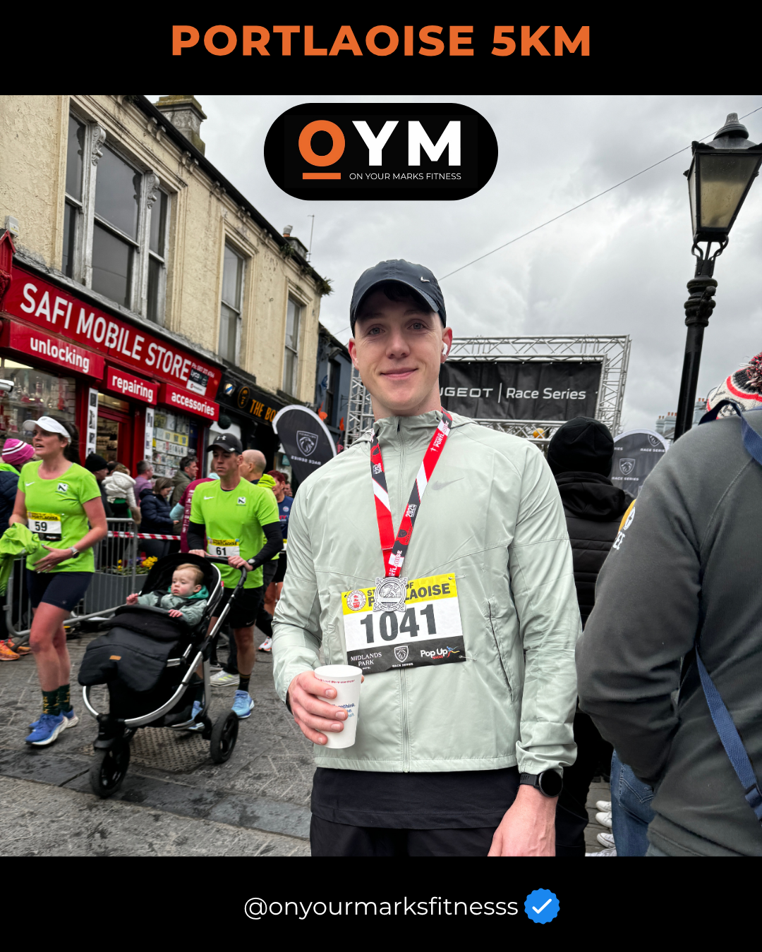 A man wearing a gray jacket and black cap at a 5K race, holding a small cup and wearing a race bib numbered 1041, with other runners and spectators in the background.