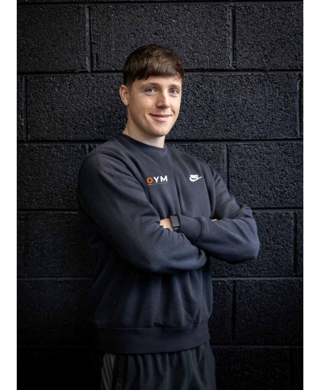 Young man with short brown hair and blue eyes standing with arms crossed, smiling, wearing a black Nike sweatshirt with OYM and Nike logos, against a black brick wall background.
