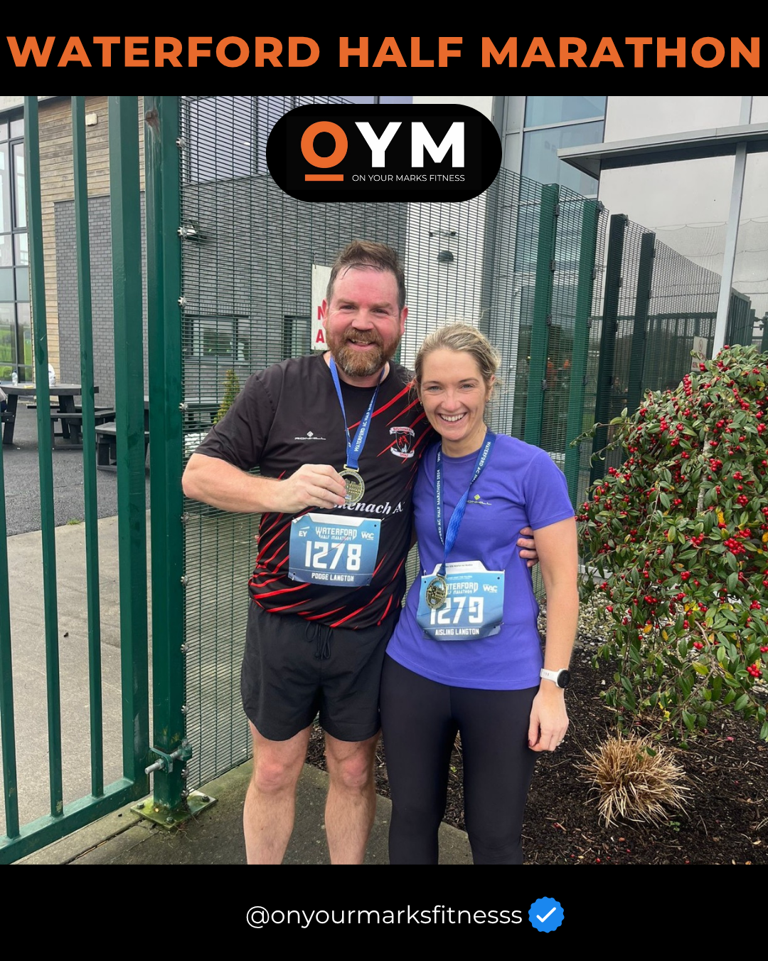 Two runners celebrating after finishing the Waterford Half Marathon, wearing medals and race bibs, standing in front of a fence with plants nearby.