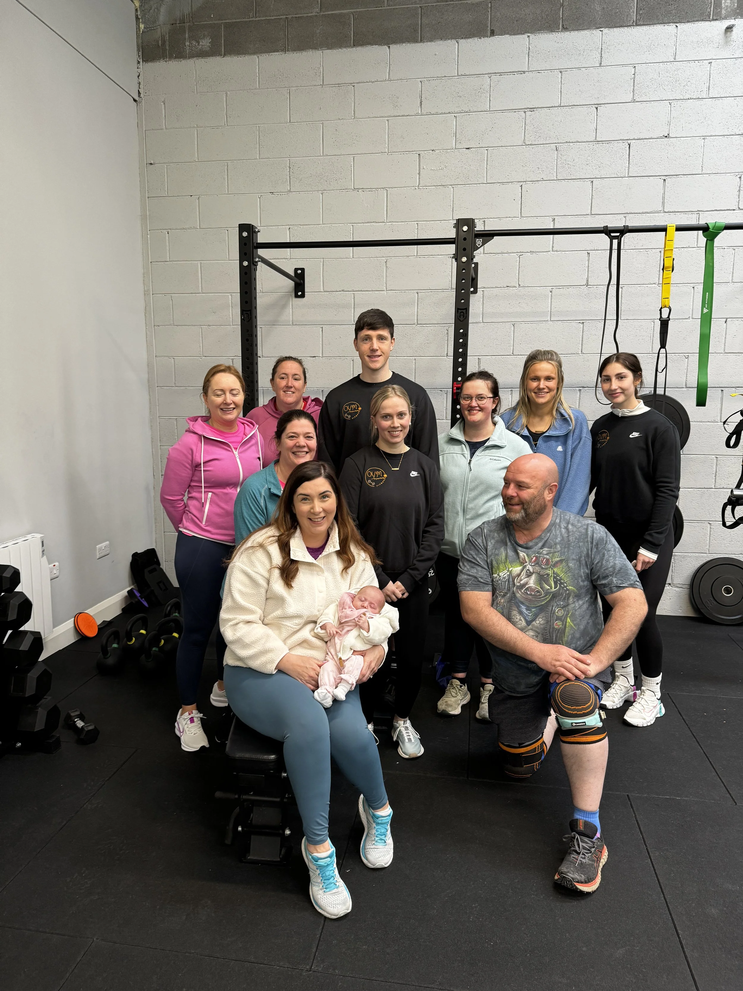 Group of people in a gym with some wearing workout clothes, a woman holding a newborn baby, and man with a prosthetic leg, posing in front of workout equipment.