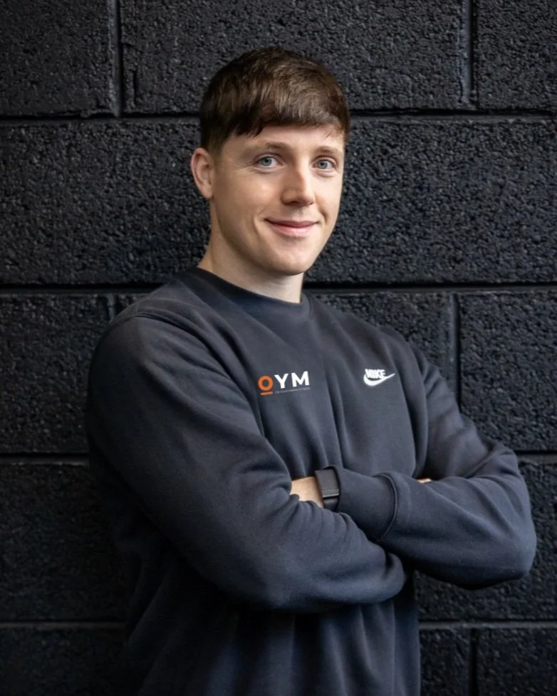 Young man with brown hair, wearing a black Nike sweatshirt with OYM logo, smiling, standing against a dark textured wall with arms crossed.