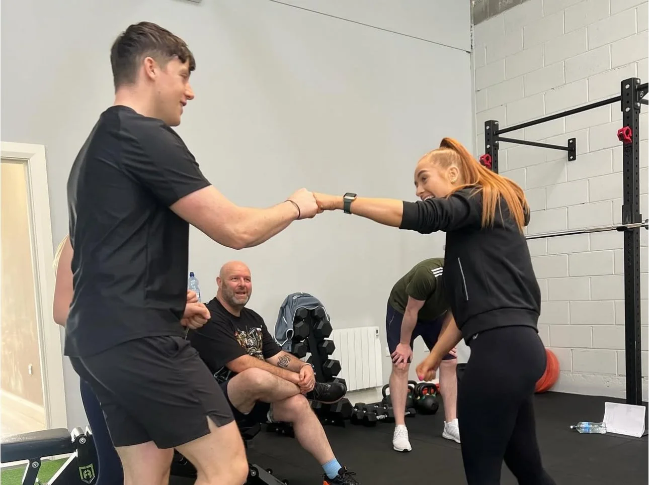 A woman in workout clothes playsfully pulls on a man's arm while others watch in a gym setting.