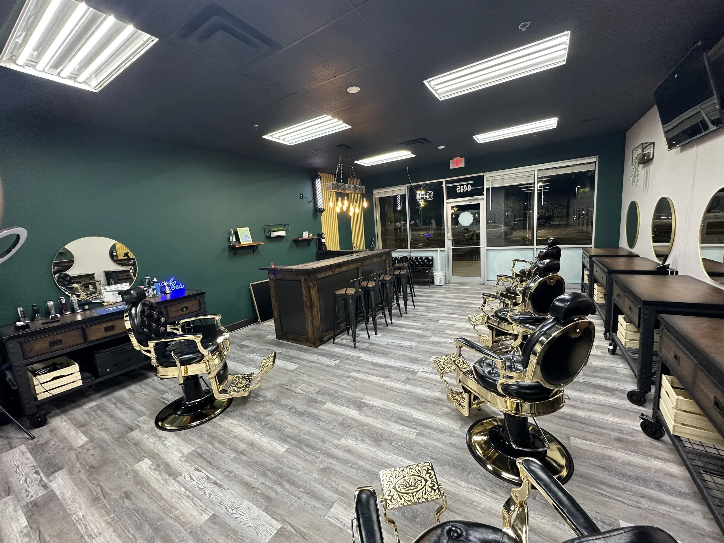 Empty barber shop interior with black and gold vintage style barber chairs, wooden flooring, dark green and white walls, large front windows, and mirrors.