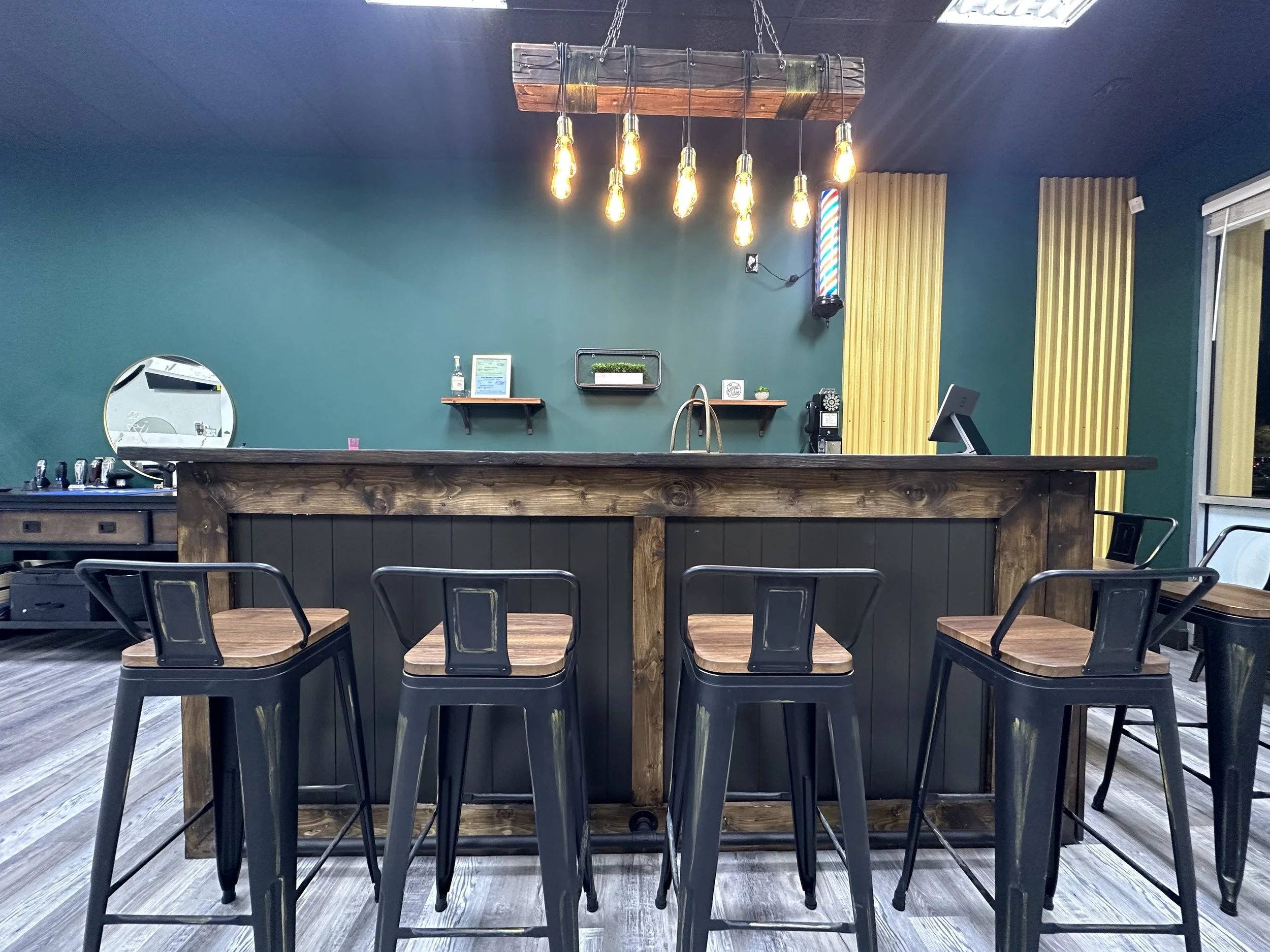 Empty bar counter with four black metal chairs, a teal wall with small shelves, hanging Edison bulbs, wooden and yellow acoustic panels, and a window with a view outside.
