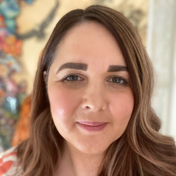 Close-up of a woman with wavy brown hair, light makeup, and a slight smile, standing indoors with blurred colorful decor in the background.