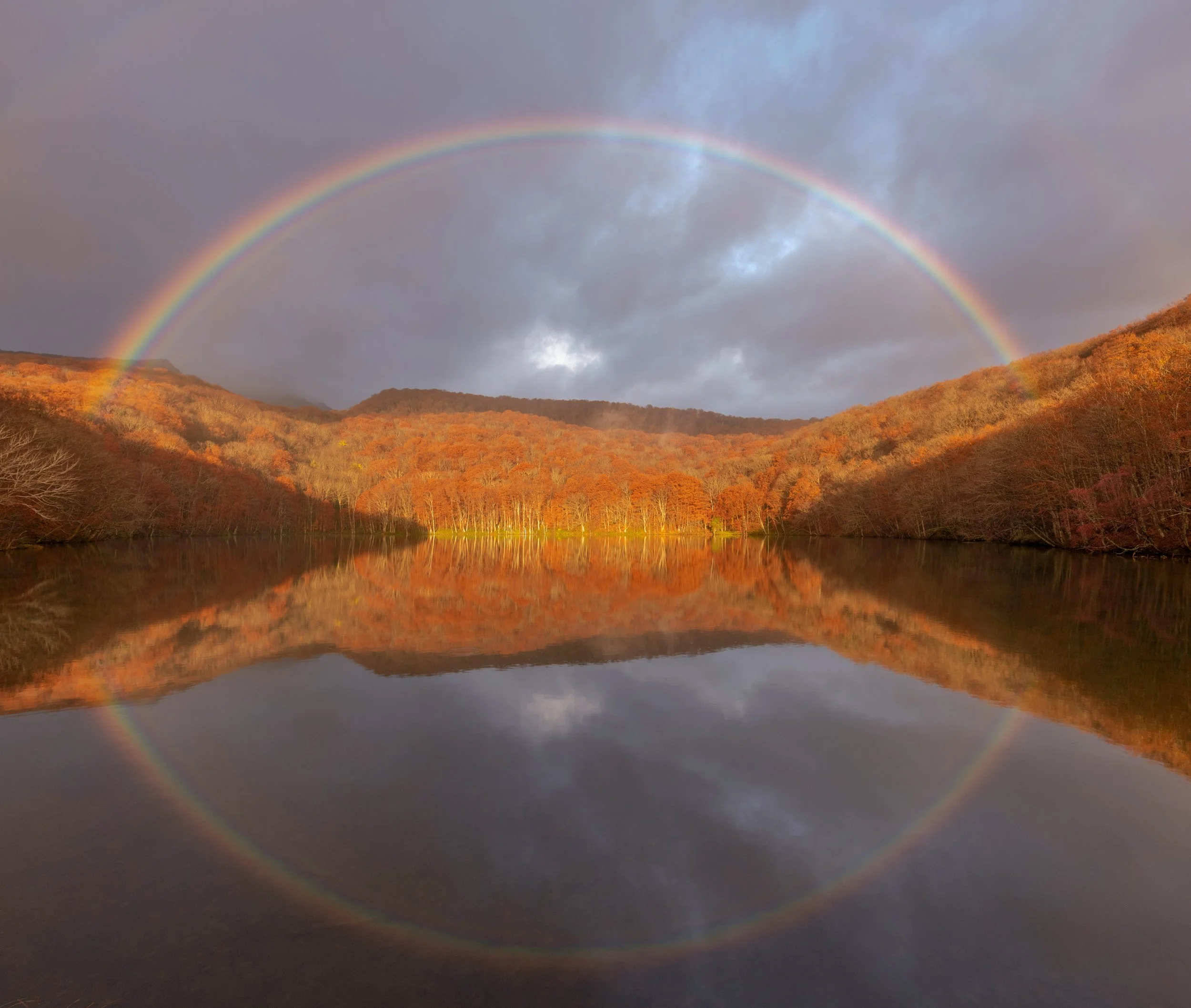 A rainbow arching over a forested mountain lake, with orange and brown trees reflecting in the water, and dark clouds in the sky.