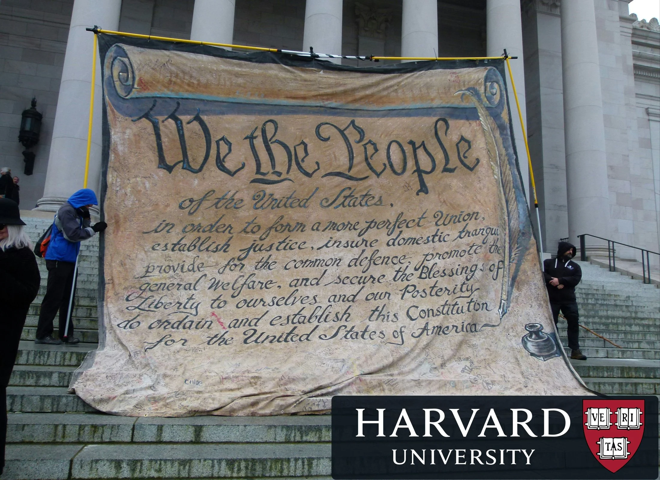 A large fabric banner depicting the preamble to the United States Constitution is displayed on the steps of a government building. Two people are holding the banner. In the bottom right corner, the Harvard University logo is overlaid on the image.