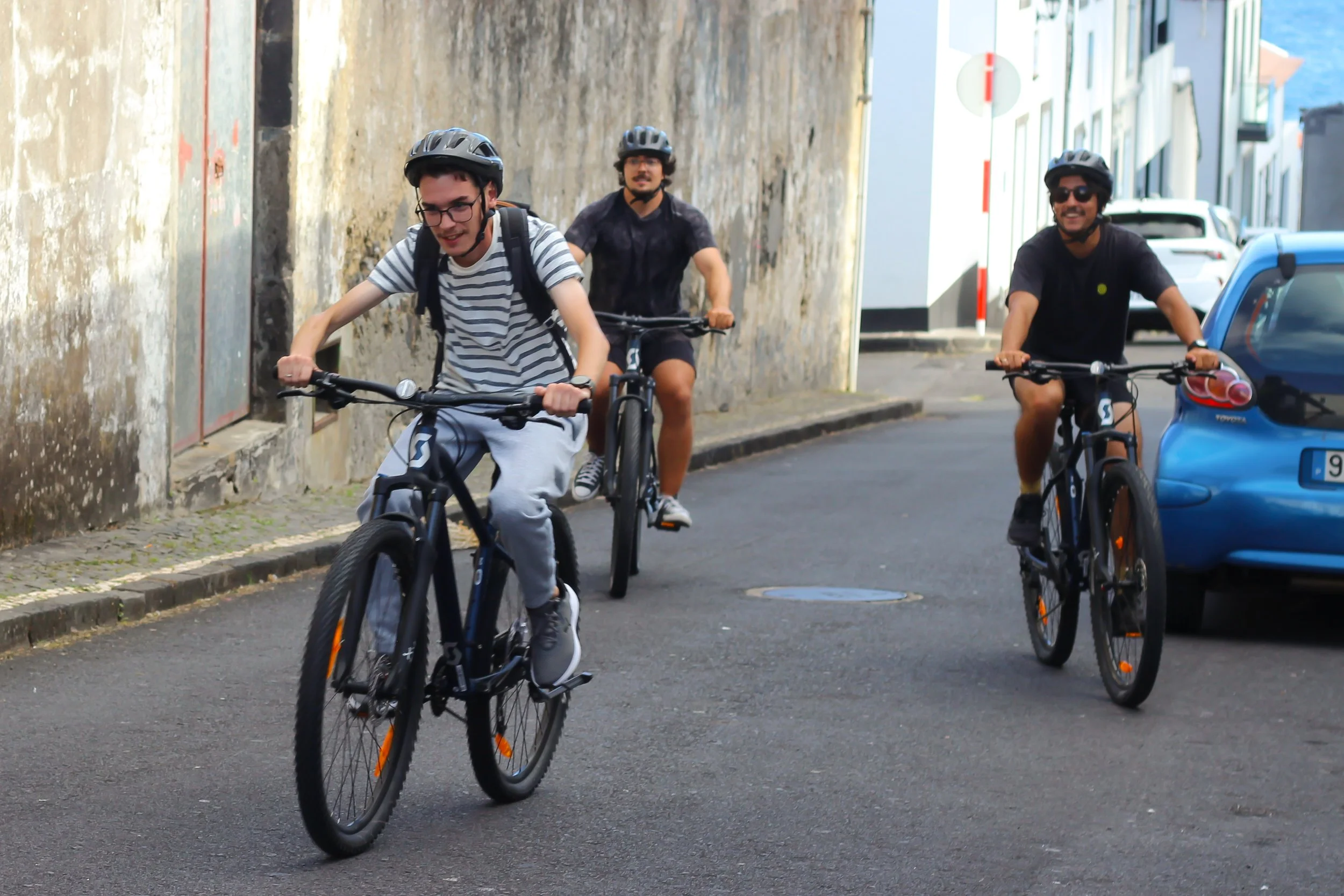 Three young men are riding bicycles on a city street, all wearing helmets and casual clothing.