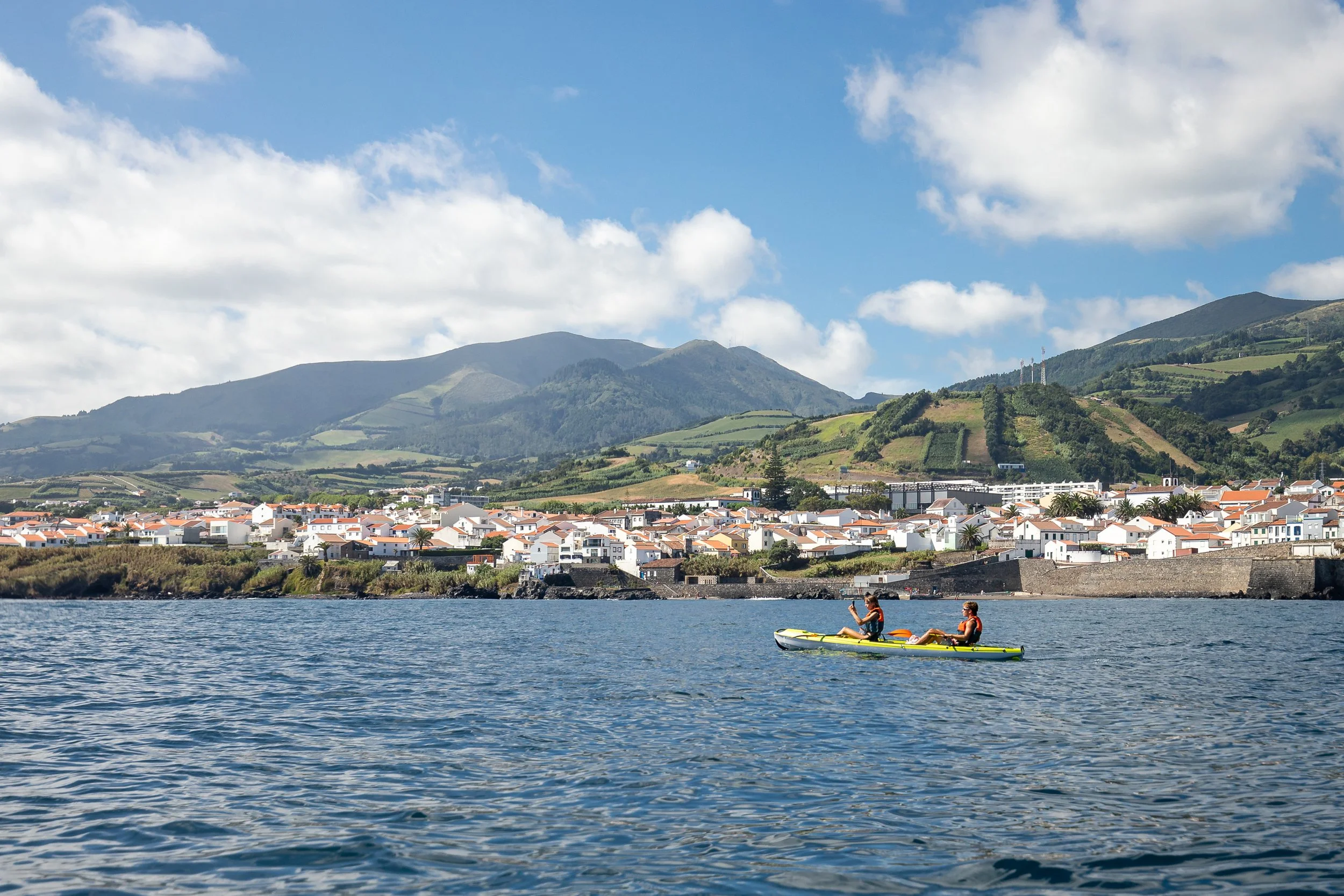 People kayaking on a body of water with a town and green hills in the background under a partly cloudy sky.