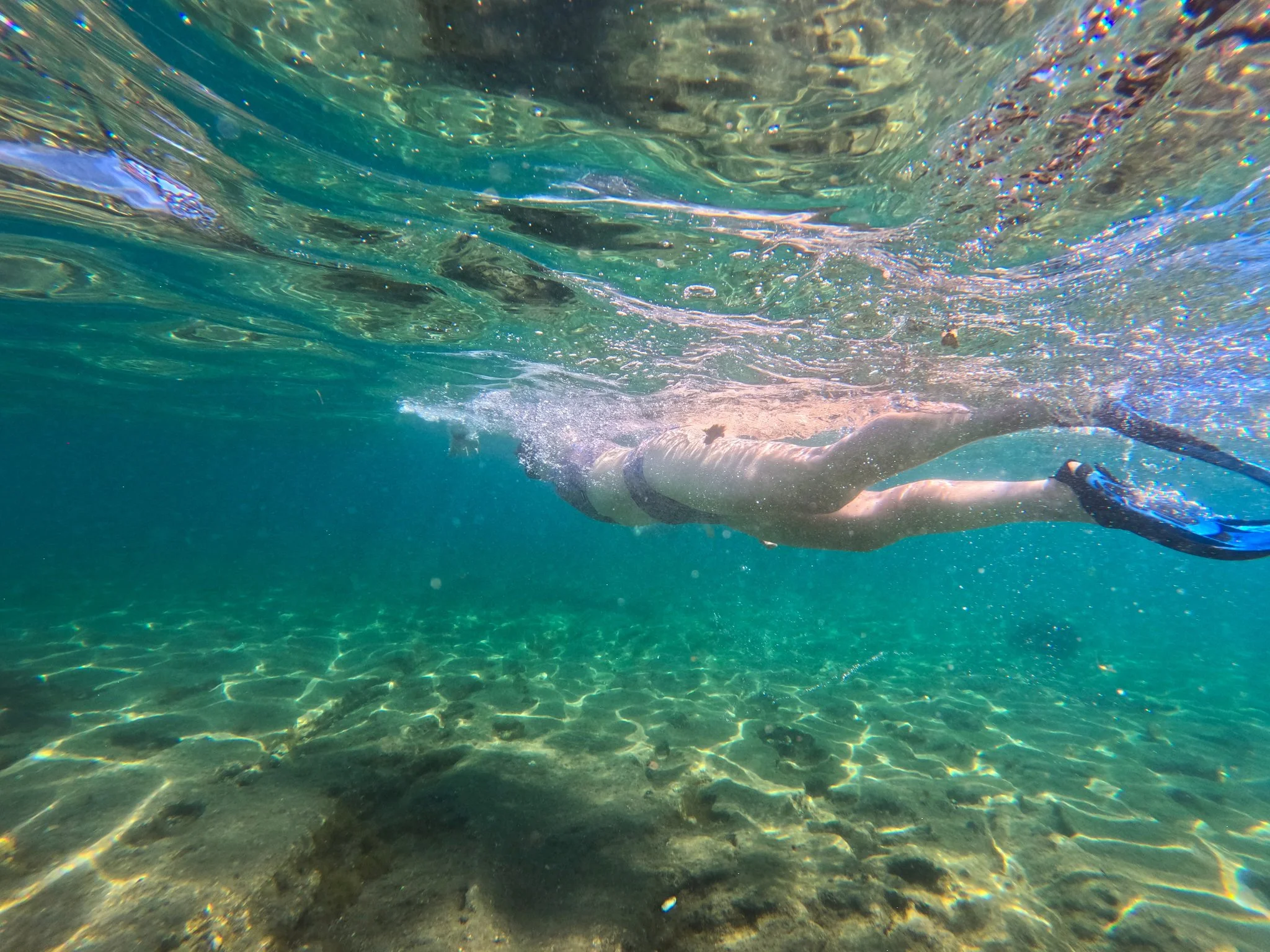 Underwater view of a person swimming in clear ocean water with sunlight reflections on the ocean floor.