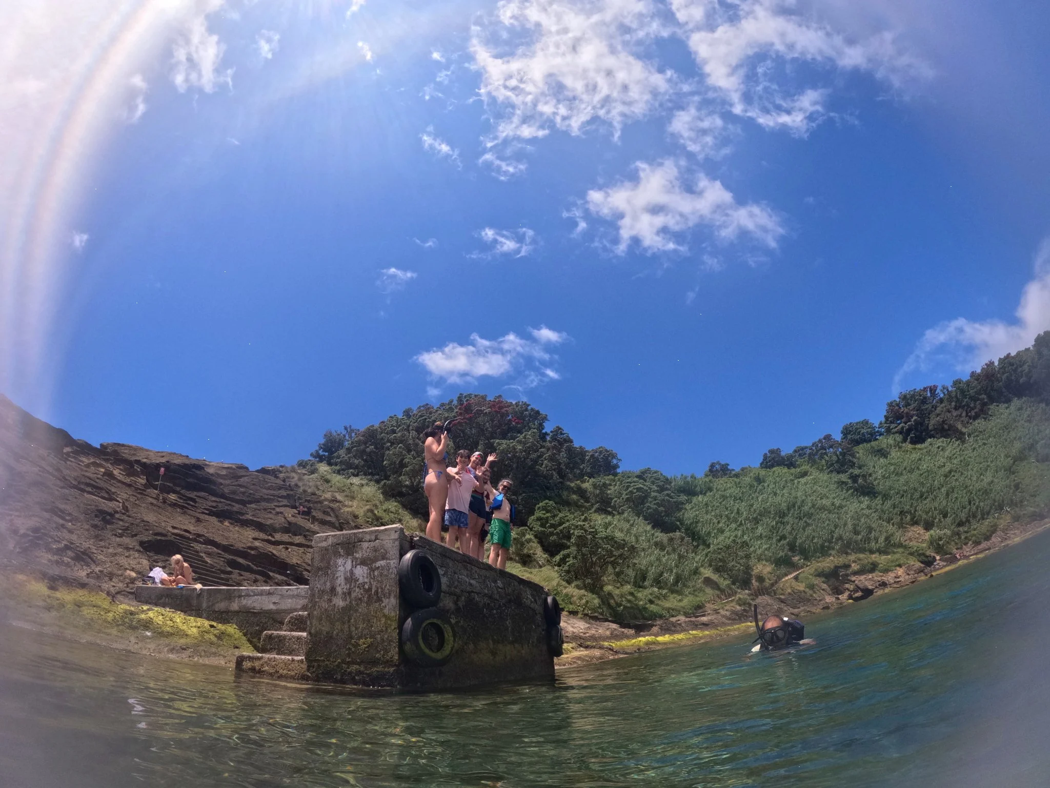 People at a riverbank with steps, trees, and a clear blue sky with some clouds, some individuals are swimming or standing near the water, and a person is lying on the steps.