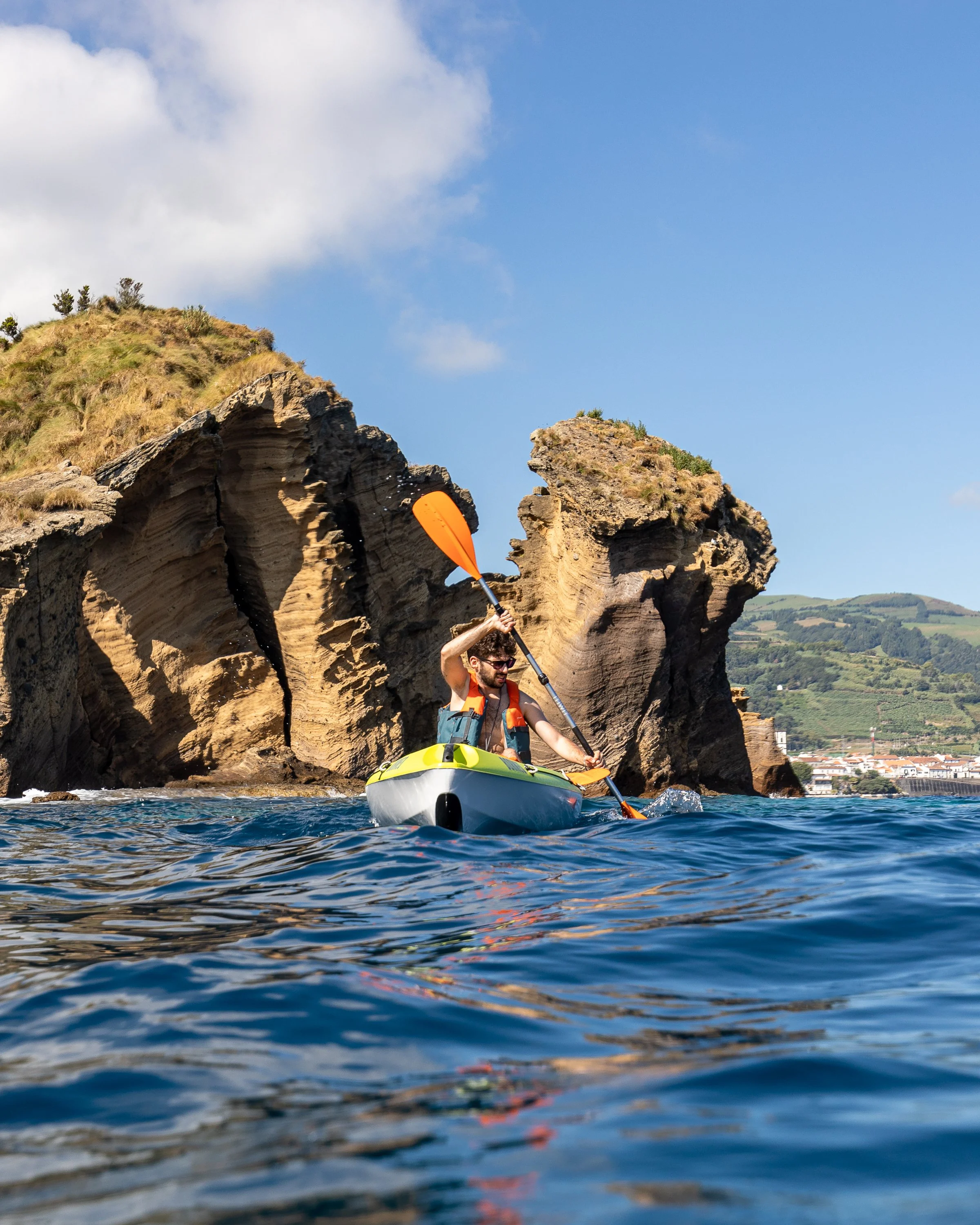Person kayaking near rocky shoreline with hills and a town in the background under a partly cloudy sky.