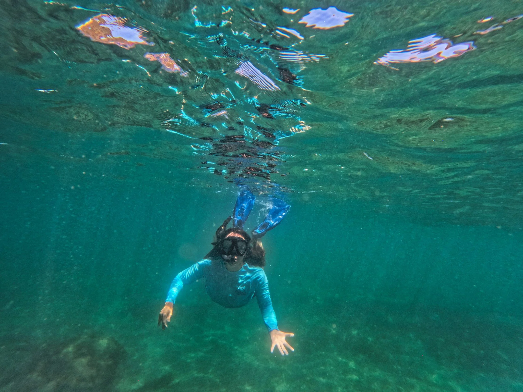 A person in a long-sleeved blue shirt and swimming trunks underwater in a pool, reaching out with one hand and swimming with fins.