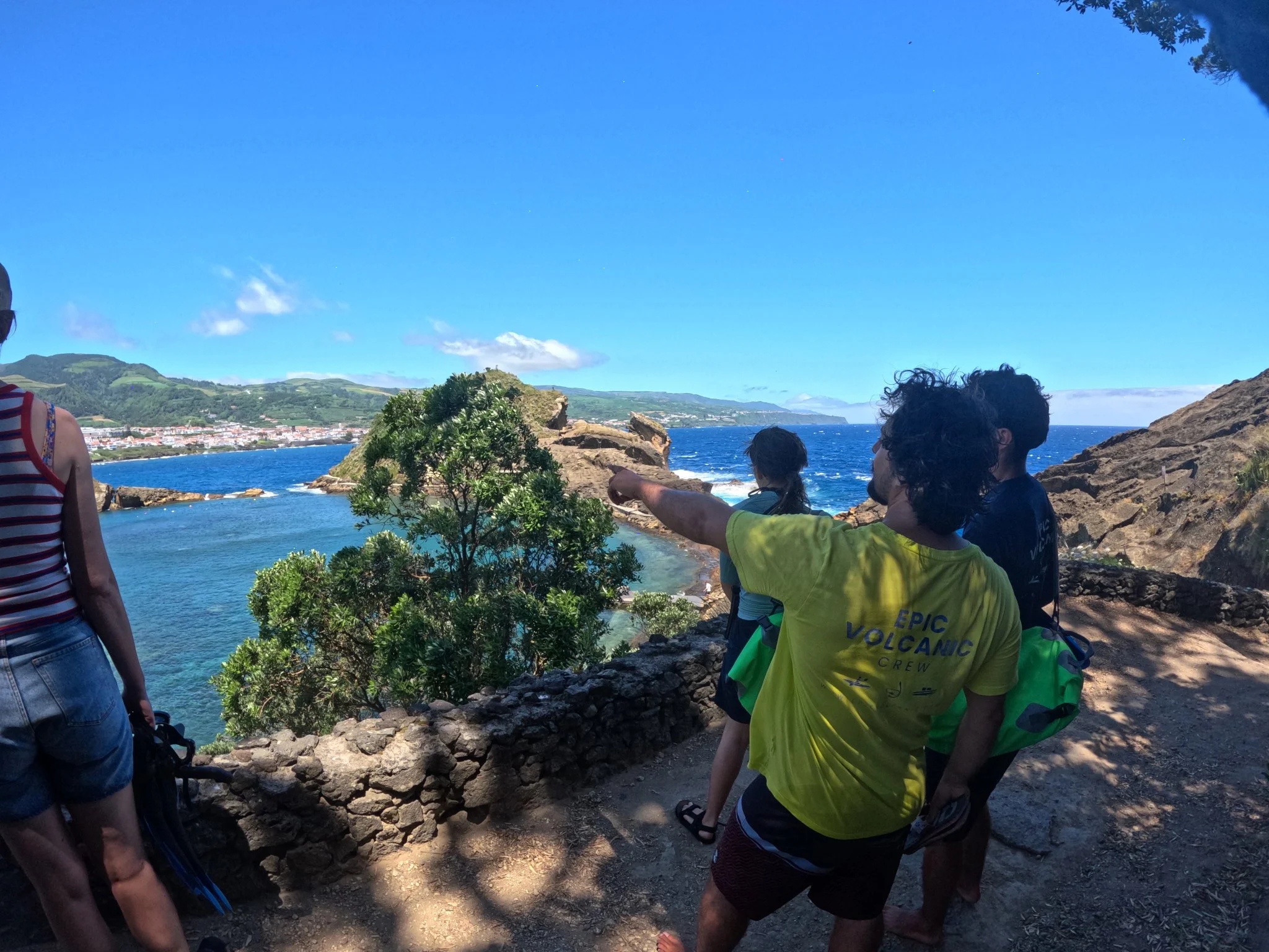 Group of people, including children and adults, standing on rocks near the ocean on a sunny day with a view of a coastal town and green hills in the background.