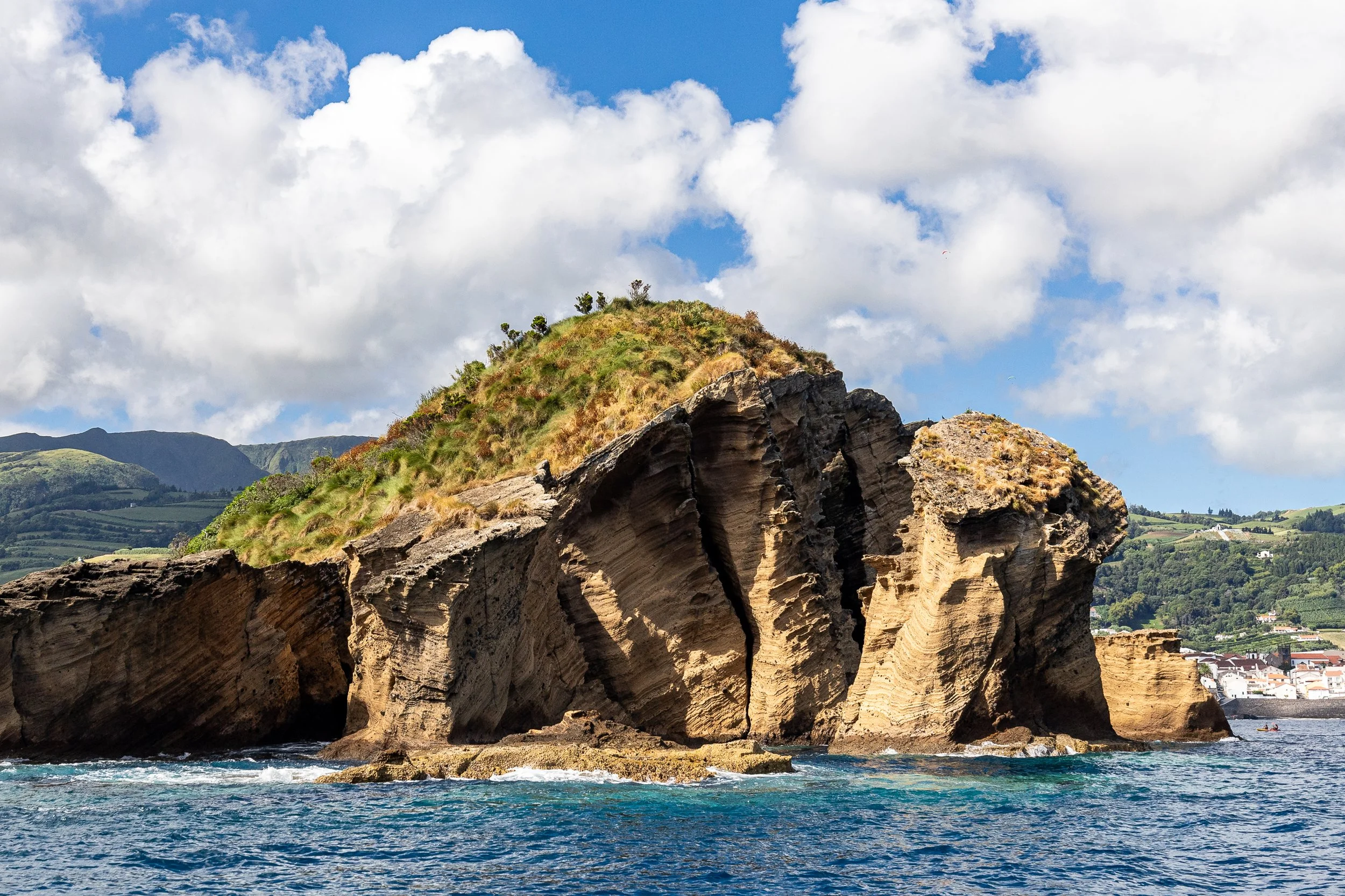 Cliffs of the Islet of Vila Franca do Campo (Princess Ring Islet) with grassy top, ocean in the foreground, and hills and buildings of Vila Franca do campo in the background under a partly cloudy blue sky.