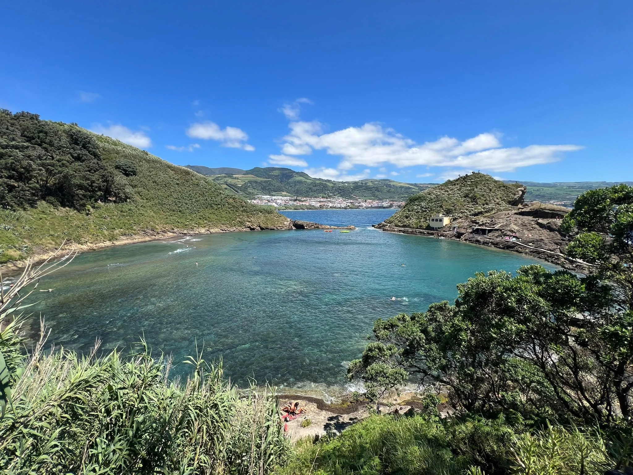 A scenic view of a bay with clear blue water, surrounded by green hills and mountains under a partly cloudy sky, with a small beach and a few people visible at the shoreline.