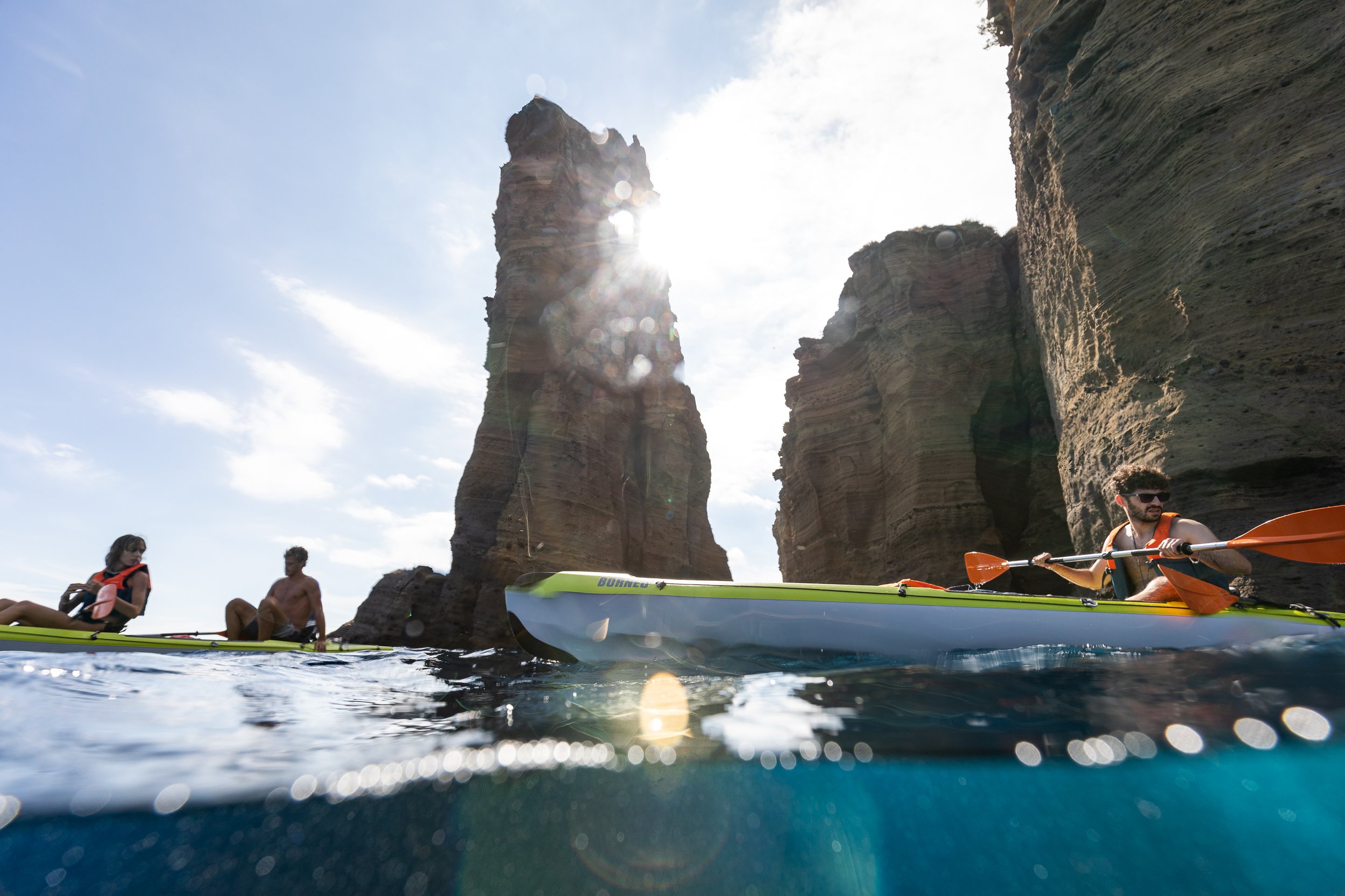 People kayaking near tall rock formations with cliffs, under a bright sky with the sun shining.