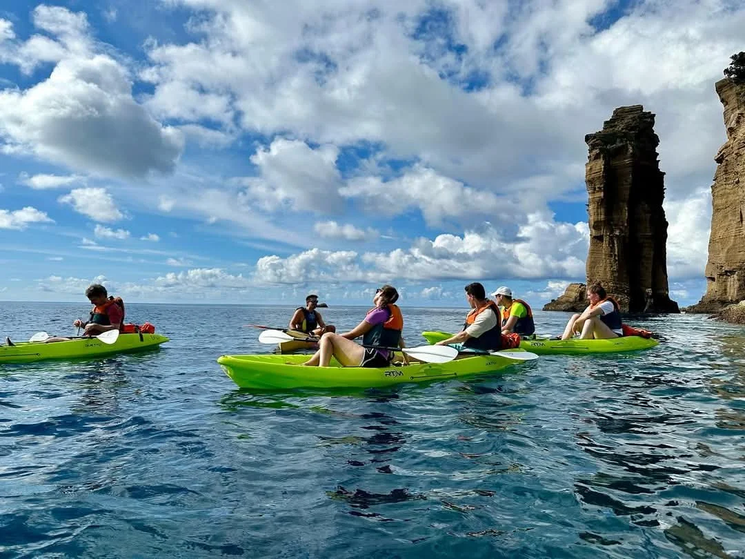 A group of six people kayaking on the ocean near rocky cliffs under a partly cloudy sky.