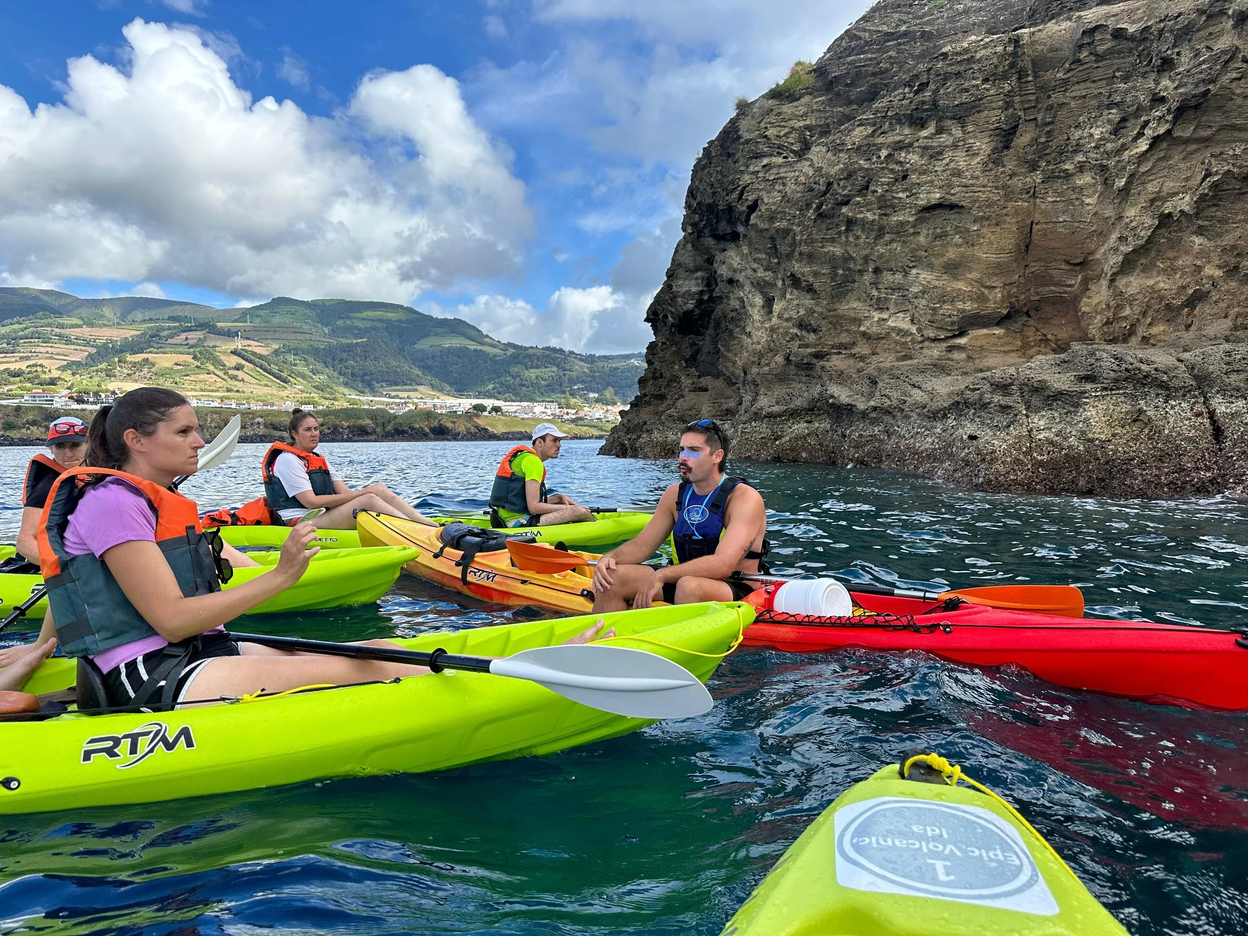 Group of people kayaking near a rocky cliff and green hills under cloudy sky.