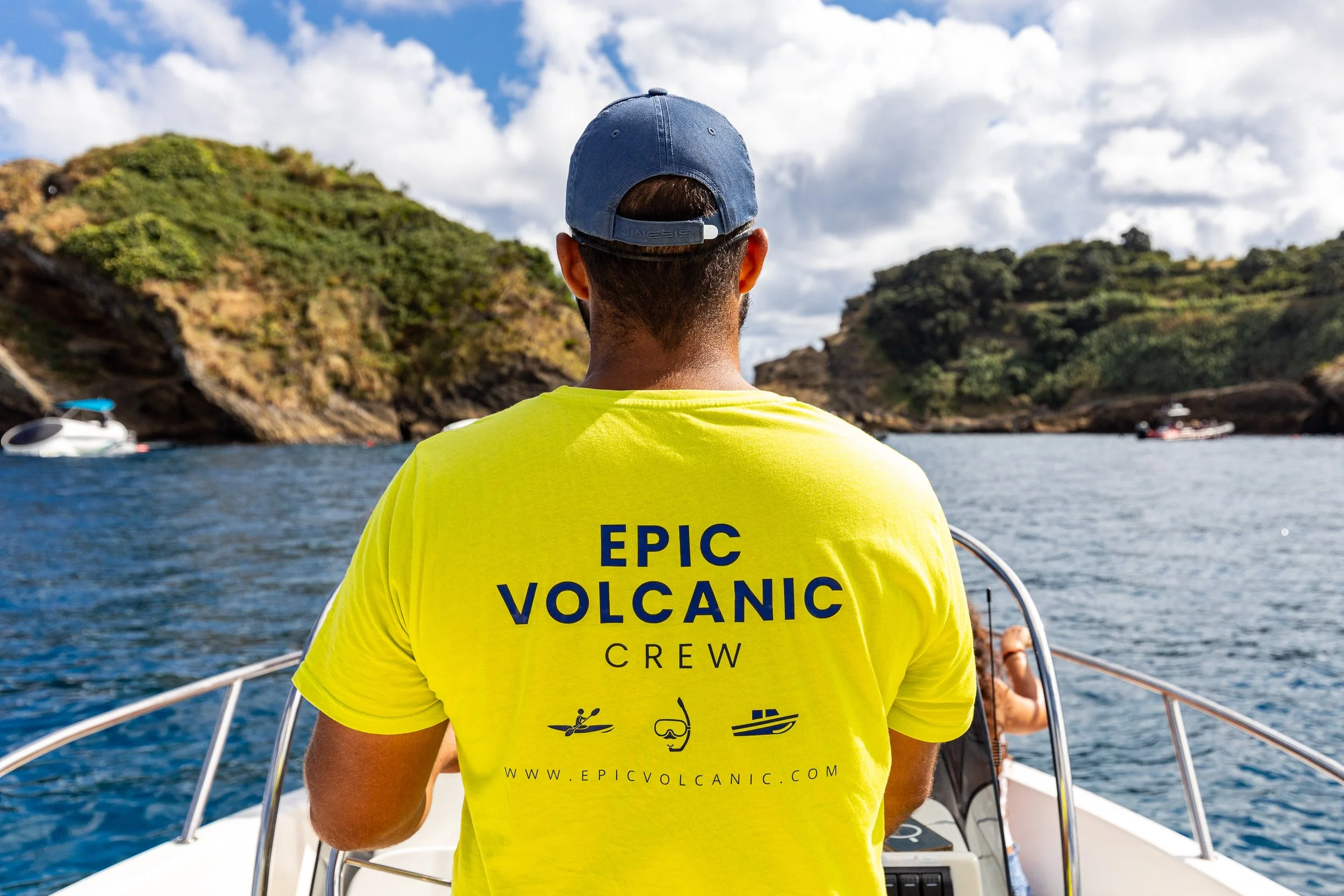 Rear view of a Epic Volcanic crew member in a yellow shirt and blue cap steering a boat in the Islet of Vila Franca do Campo (Princess Ring Islet).