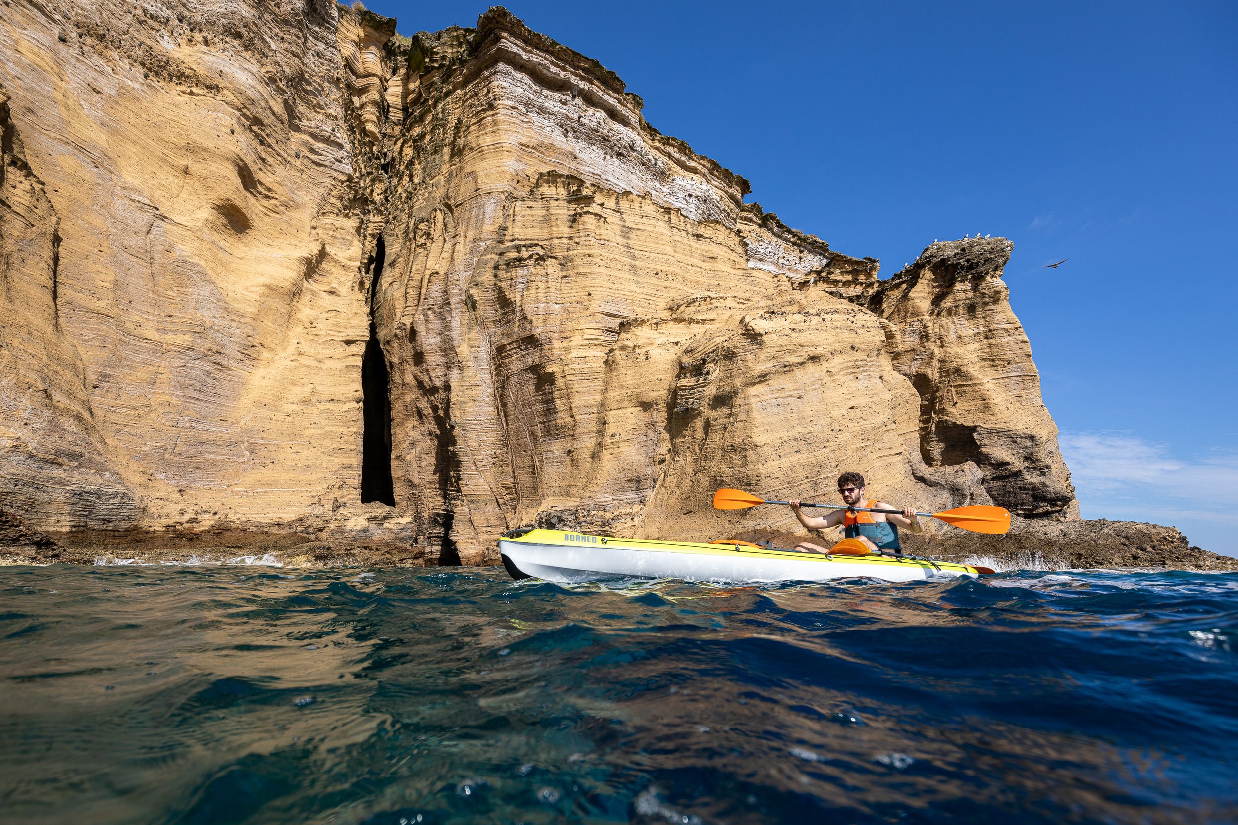 A person kayaking near a large rock formation with layered sedimentary strata, under a clear blue sky.