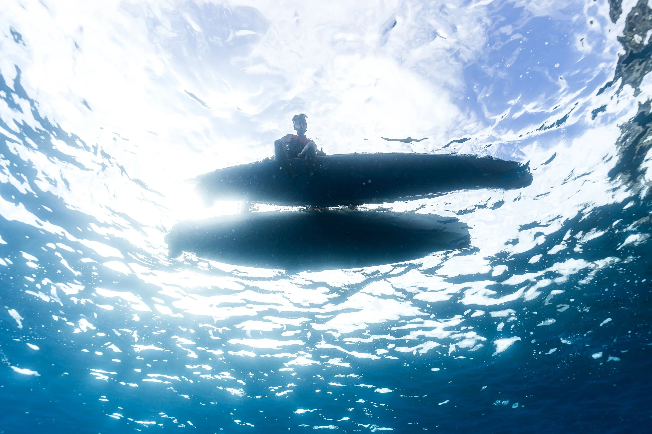 View from underwater looking up at a small boat and a person sitting in it, surrounded by the ocean's surface and waves.