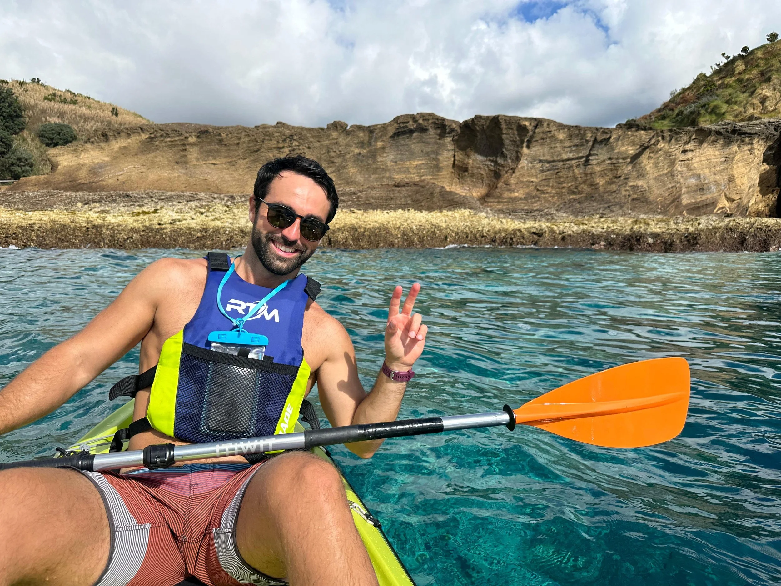 A man kayaking in clear blue water near rocky cliffs. He is smiling, wearing sunglasses and a blue life vest, and making a peace sign with his hand.