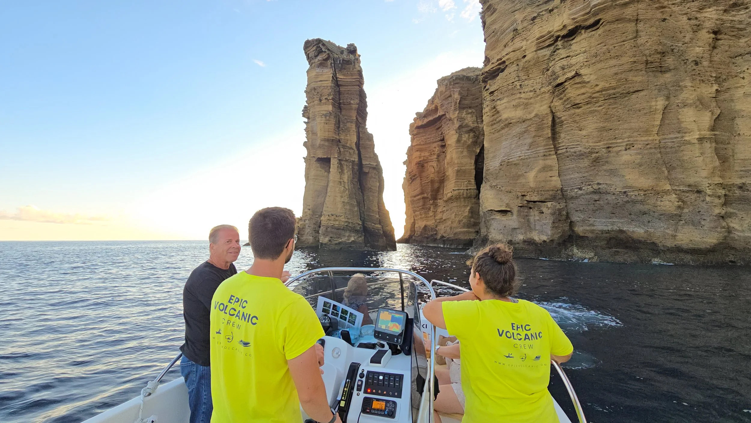 People on a boat viewing tall cliffs and sea stacks during sunset. Two people wear bright yellow shirts labeled "Epic Volcanic Crew".