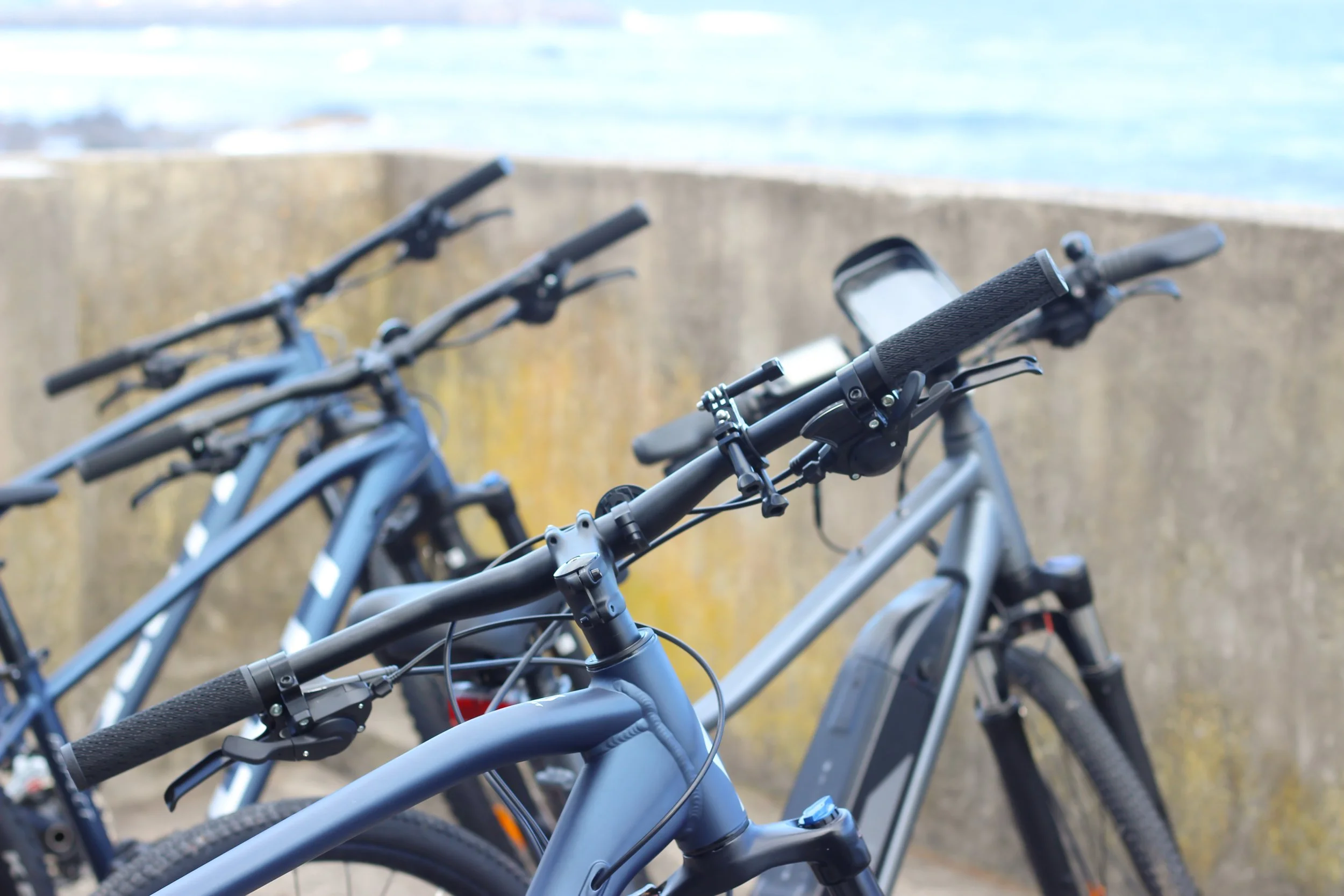 Close-up of multiple bicycles with handlebars and frames visible, parked outdoors near a concrete wall with water in the background.