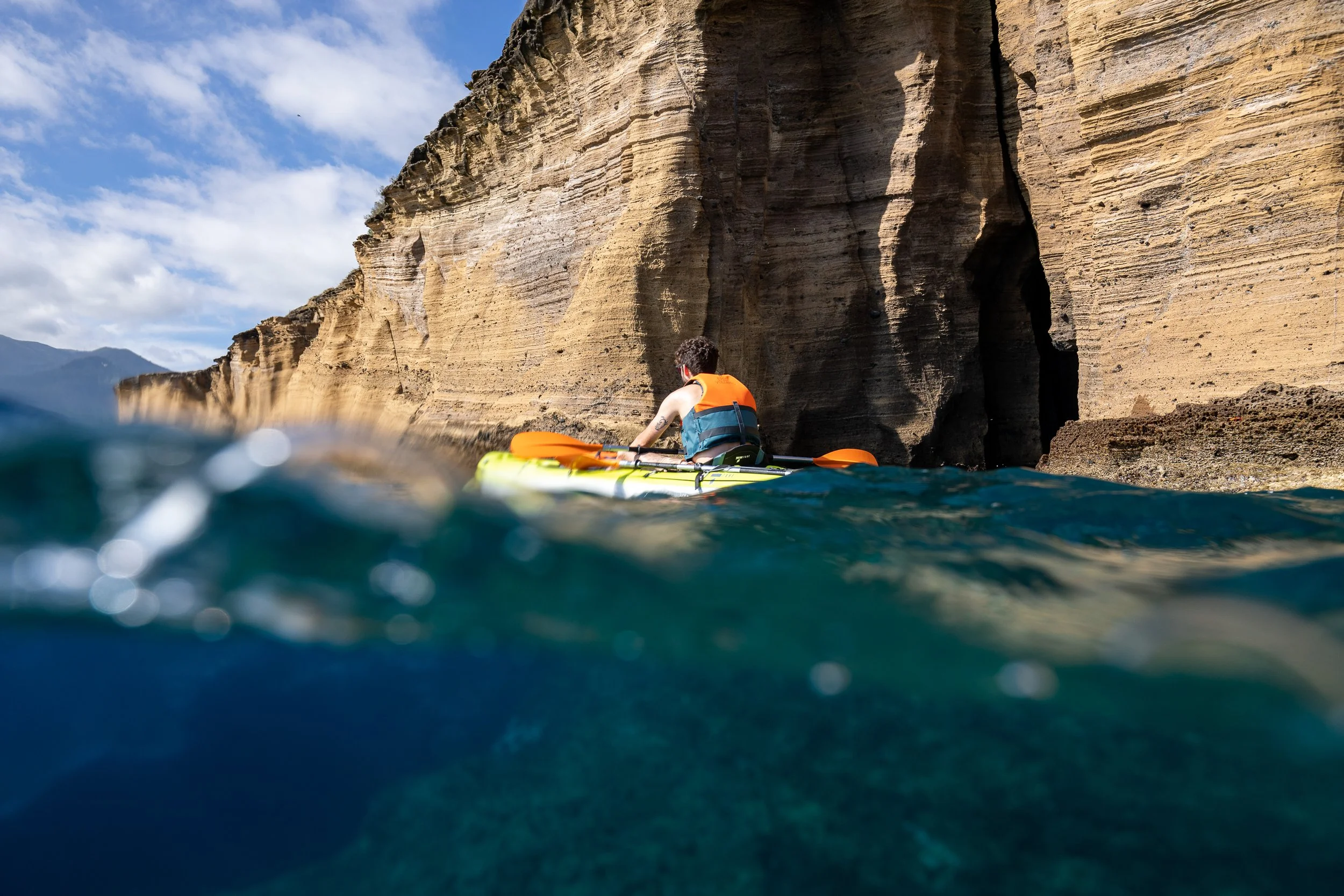 A person kayaking in the ocean near a rocky cliff, with the shot taken partially underwater showing the water's surface and the kayak.