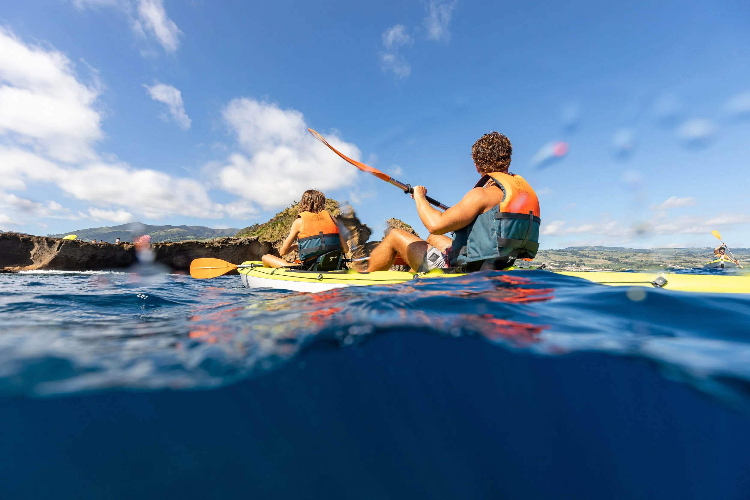 People kayaking on a clear lake with rocky shoreline and rolling hills in the background, under a partly cloudy sky.