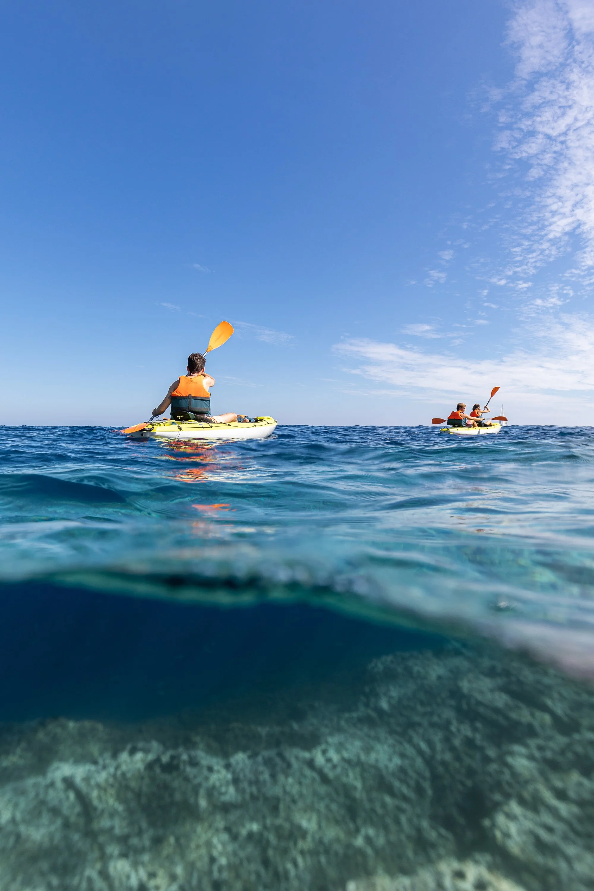 People kayaking on the ocean under a partly cloudy sky, with the photo taken from beneath the water surface showing rocks beneath.