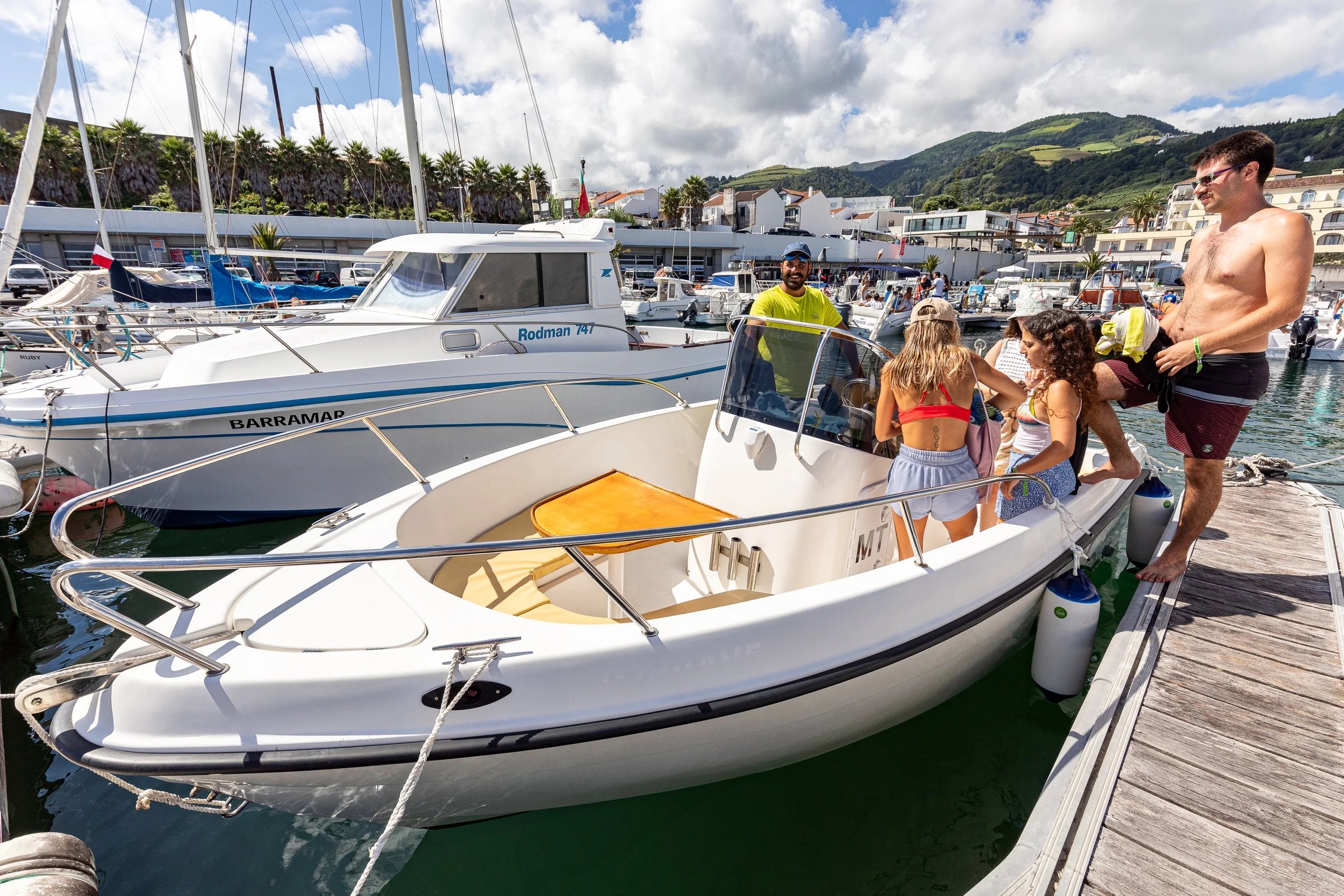 People on a dock preparing boats at a marina with mountains and buildings in the background on a sunny day.
