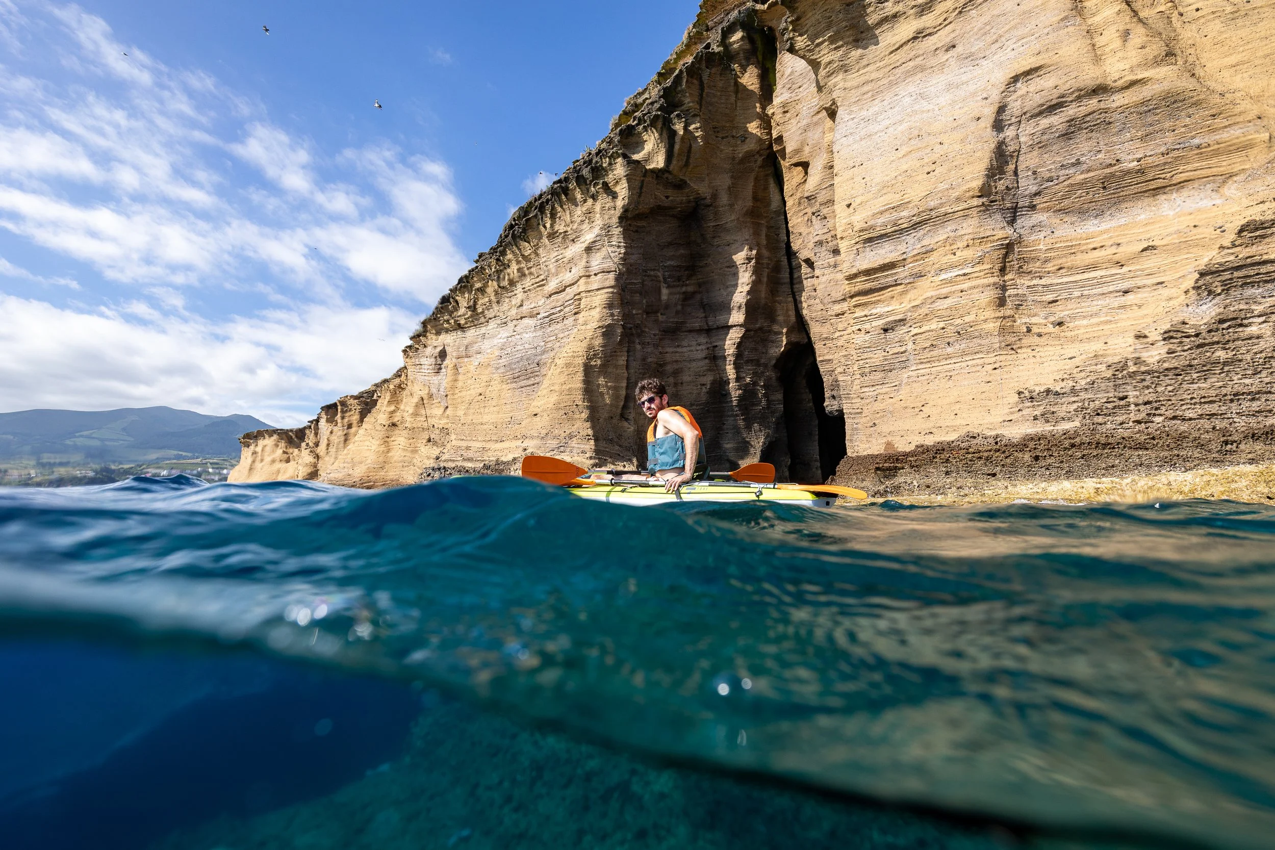A man kayaking near rocky cliffs with a mountain and blue sky in the background.