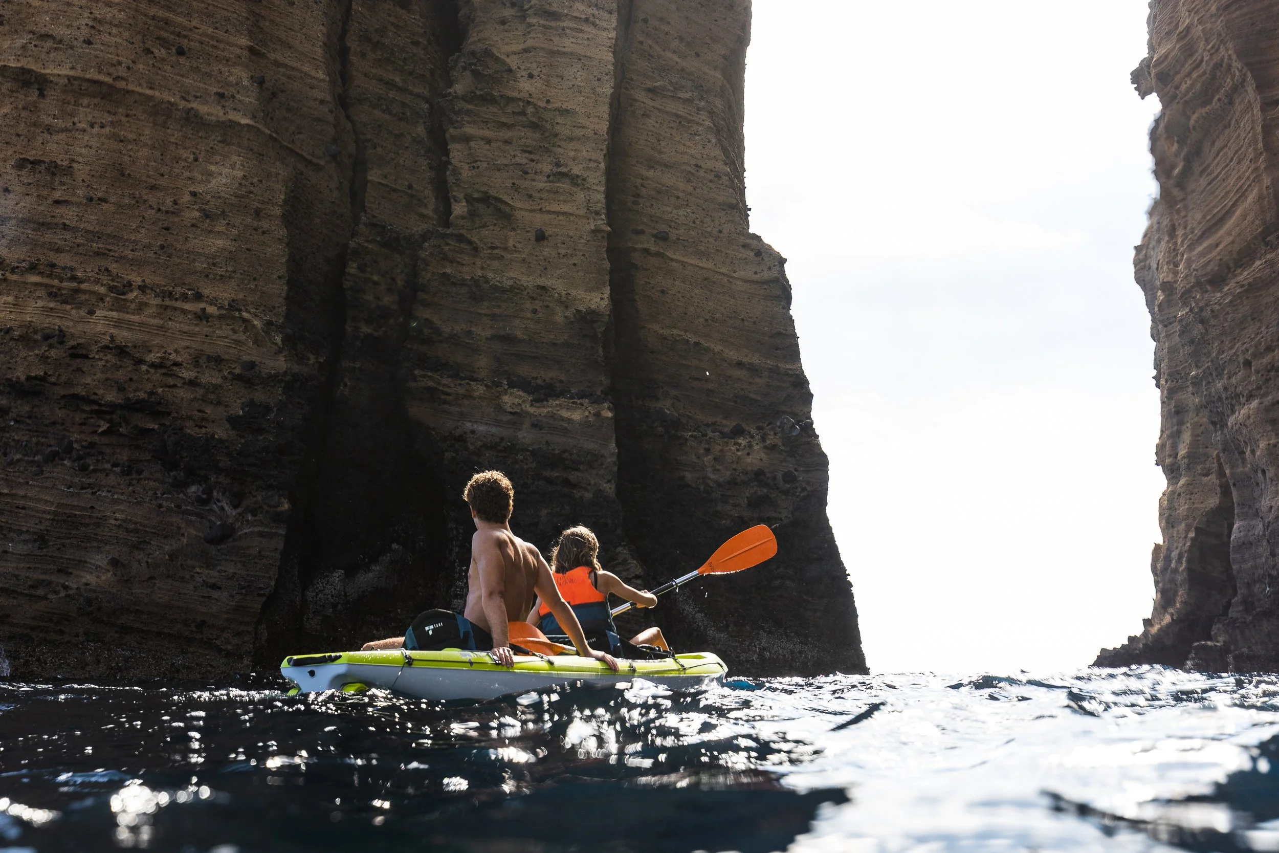 People kayaking in a narrow canyon with tall, layered rock walls on either side, and a partly cloudy sky overhead.