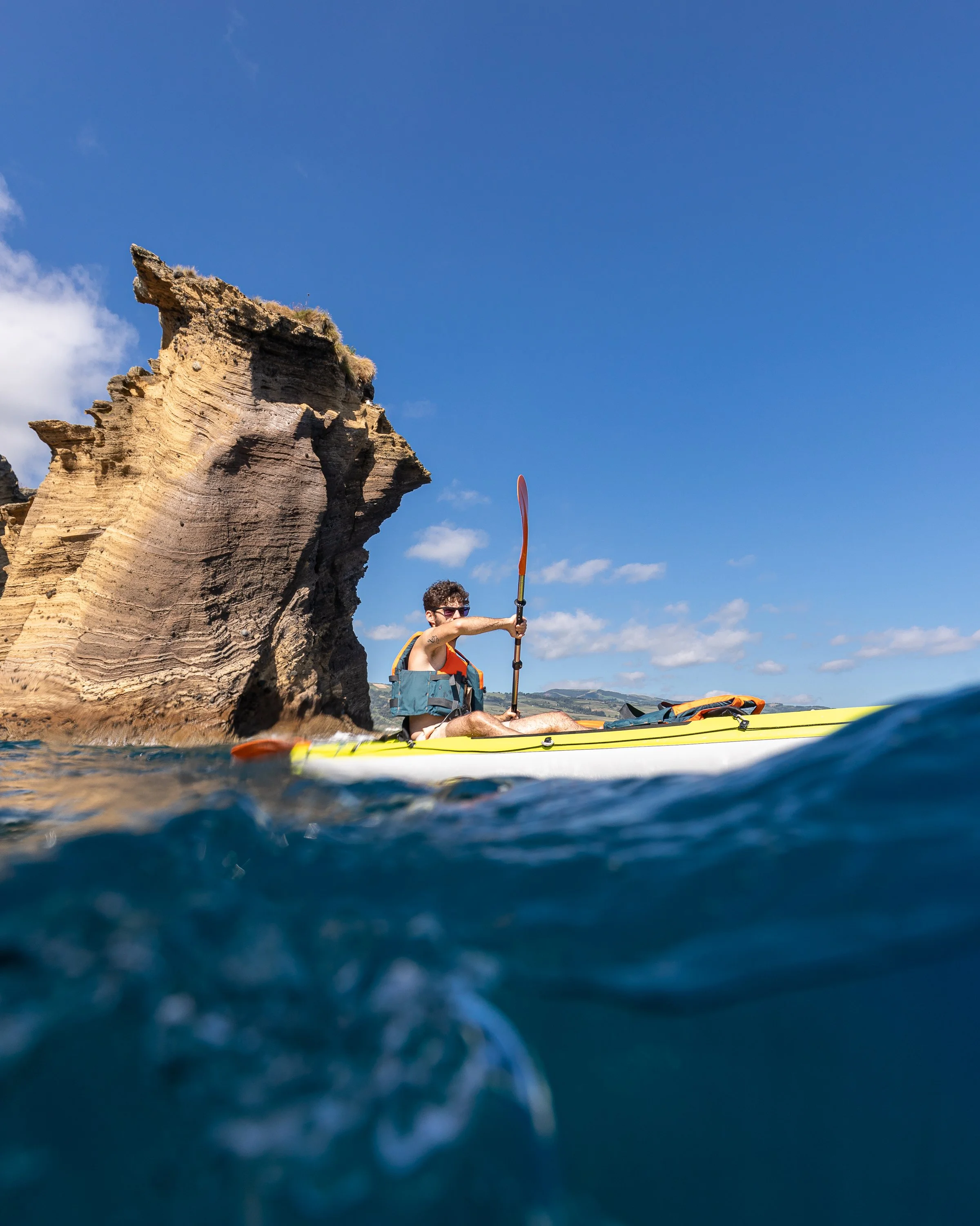 Person kayaking near a large rock formation in the Islet of Vila Franca do Campo on a clear day with blue sky and some clouds, with a split view of the ocean