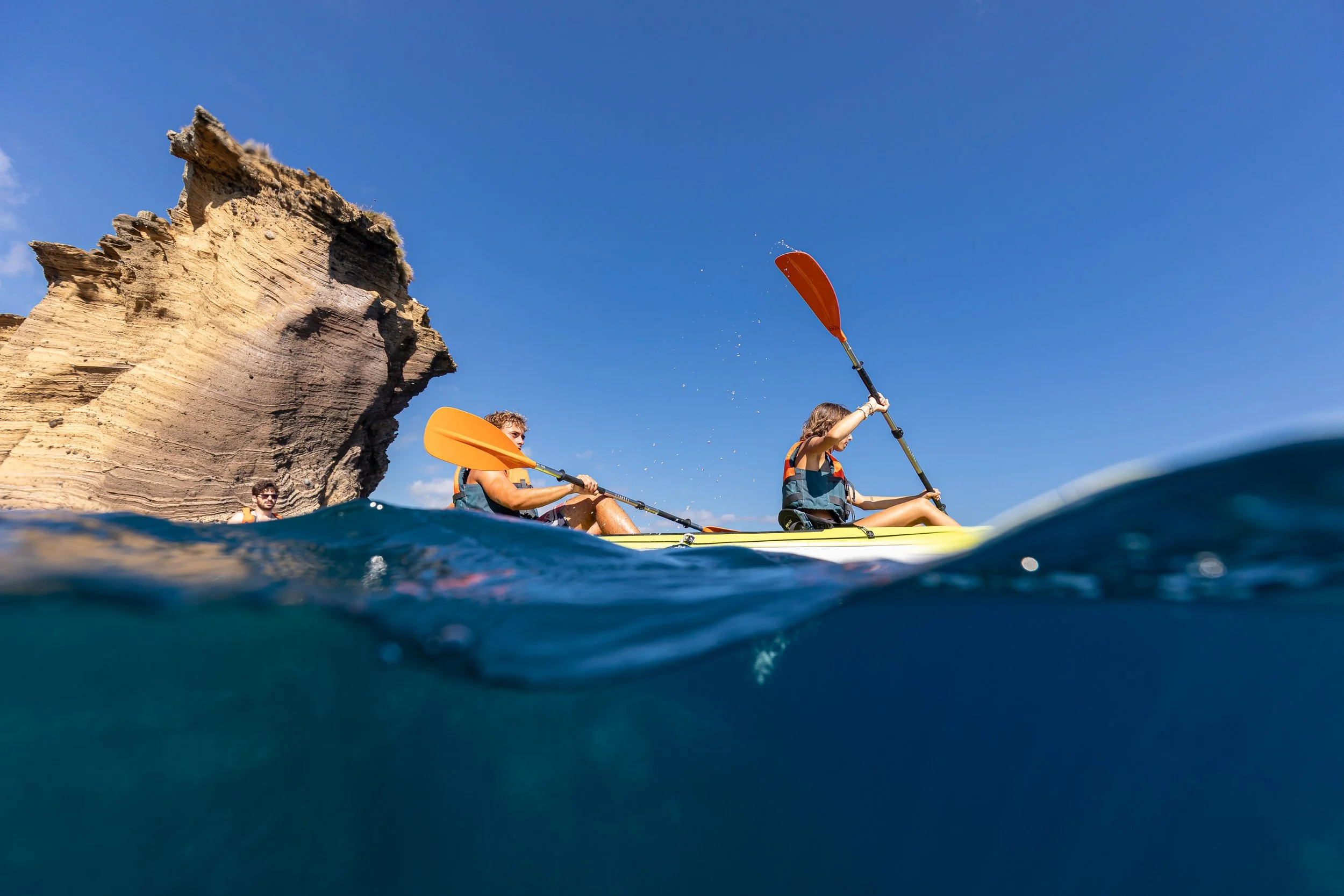 People kayaking in the water near a large, weathered rock formation under a clear blue sky.
