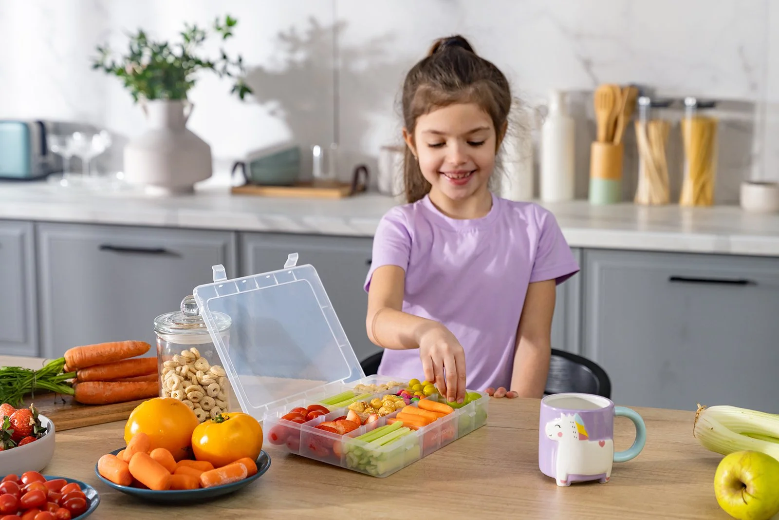 A young girl in a purple shirt packing snack foods into a lunchbox on a kitchen table. The table has fresh vegetables, berries, and a mug with a unicorn on it.