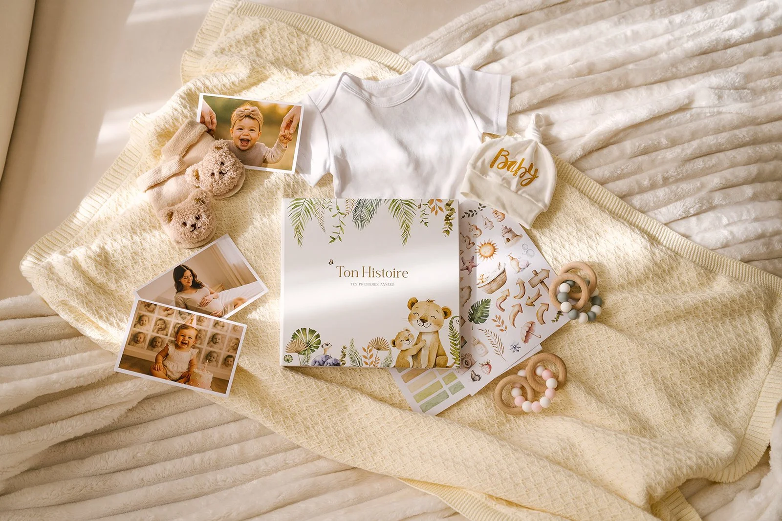 Collection of baby items including photos of a smiling toddler, a white baby hat with 'Baby' written, a children's book titled 'Ton Histoire,' plush shoes, a color palette, and teething rings, all arranged on a cream-colored textured blanket.