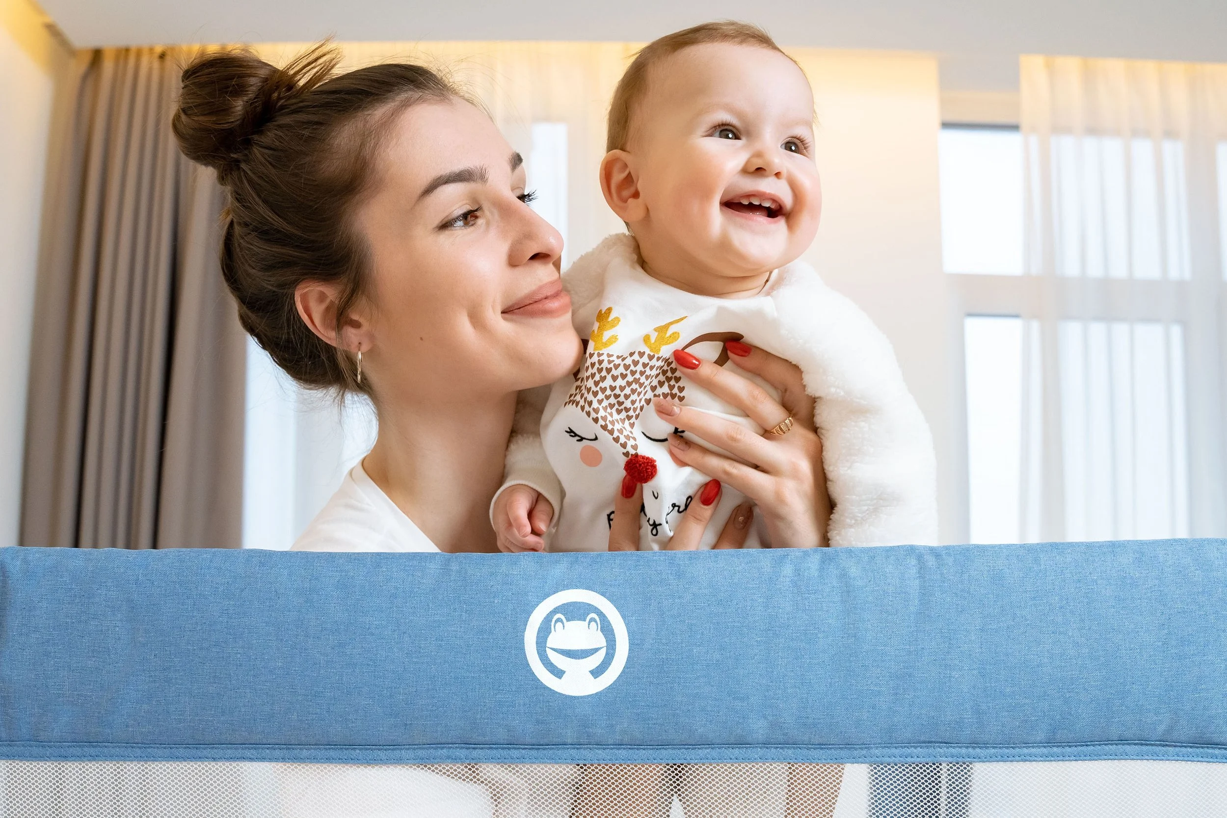 A woman with brown hair tied up in a bun holding a smiling baby girl with light hair in a white sweater with reindeer design, in a bright room with large windows and beige curtains.