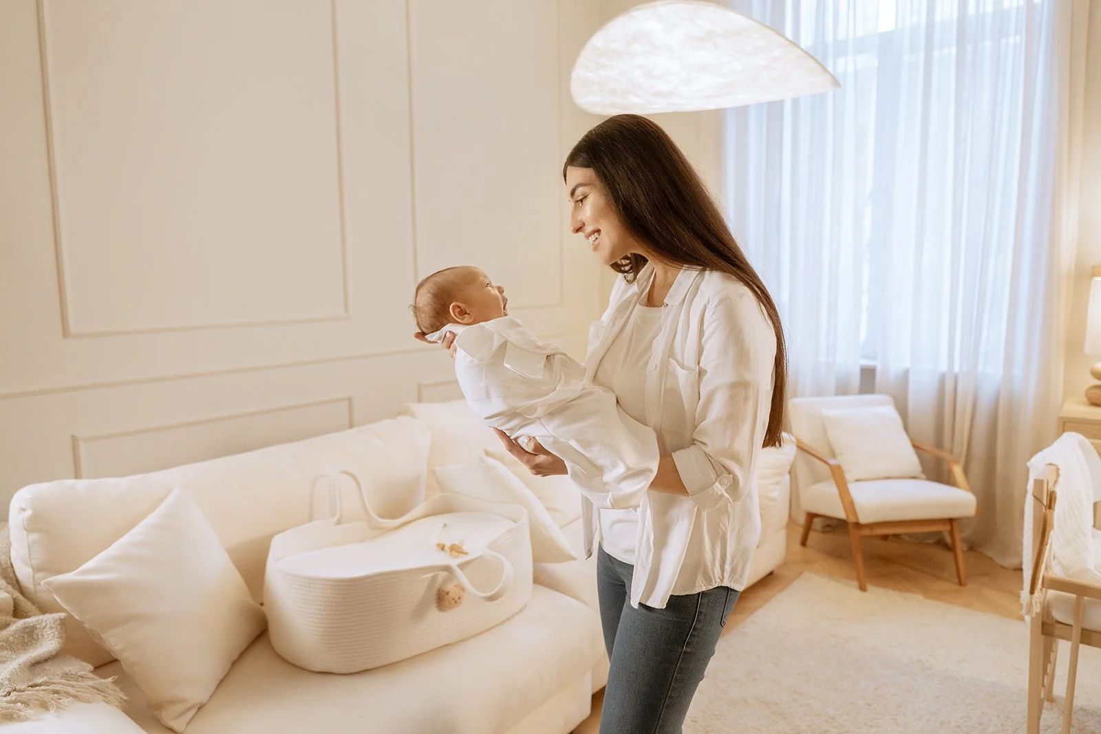 A woman holding a newborn baby inside a bright, cozy living room with white walls, a white sofa, and curtains.