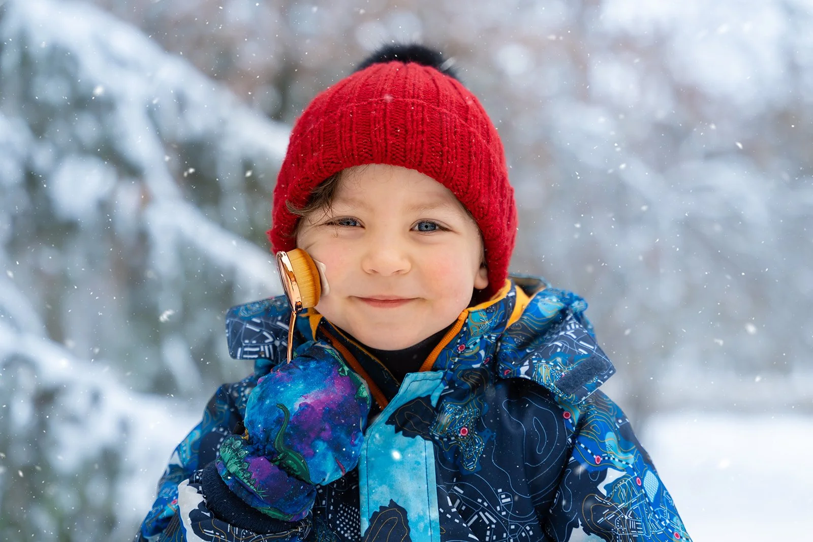 A young boy in winter clothing, including a red knit hat and a colorful jacket, using a magnifying glass to look at snow in a snowy outdoor setting.