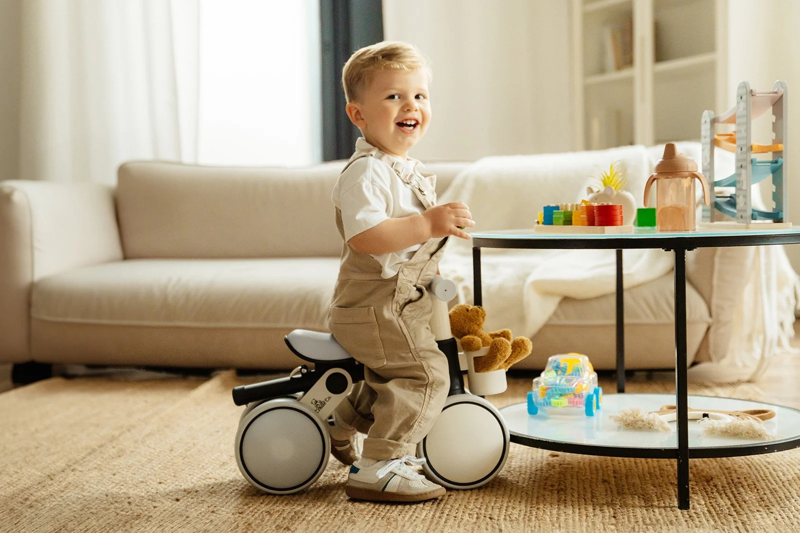 A young boy smiling while sitting on a toy bicycle in a living room, with a sofa, a window, and a table with toys in the background.